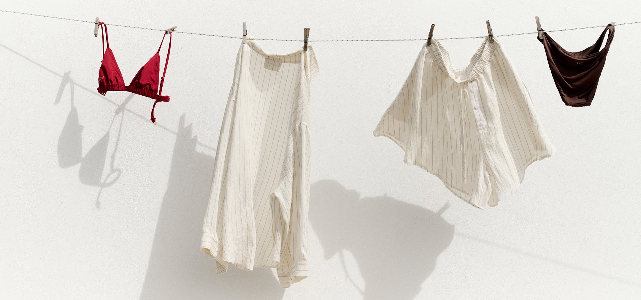 Leichte Sommerkleidung und Bikiniteile hängen an einer Wäscheleine vor weißer Wand in der Sonne.
