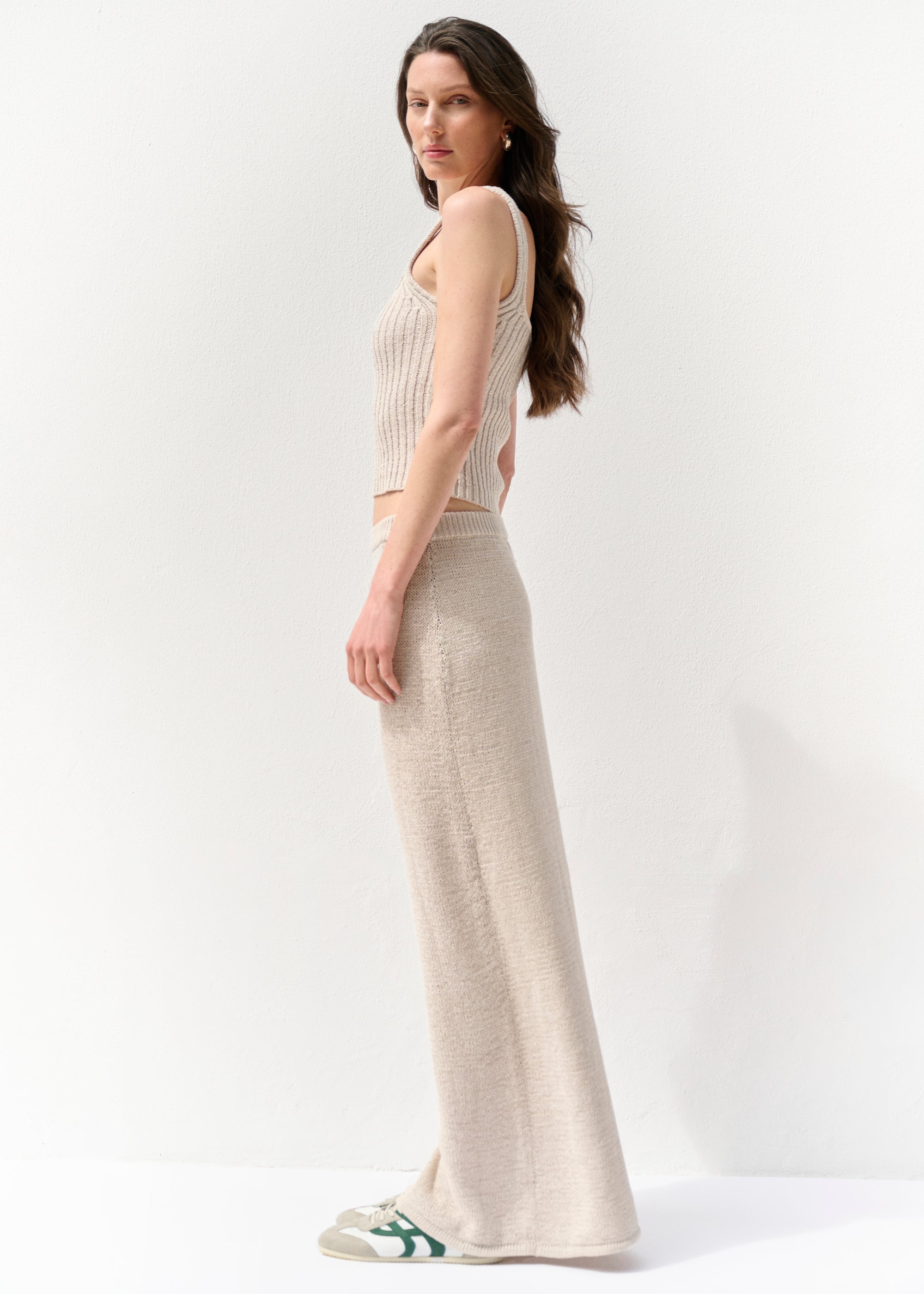 Frau im seitlichen Profil mit cremefarbenem, geripptem Stricktop mit breiten Trägern und passendem langen Rock, dazu beigefarbene Sneakers mit grünem Detail. Vor hellem Hintergrund.