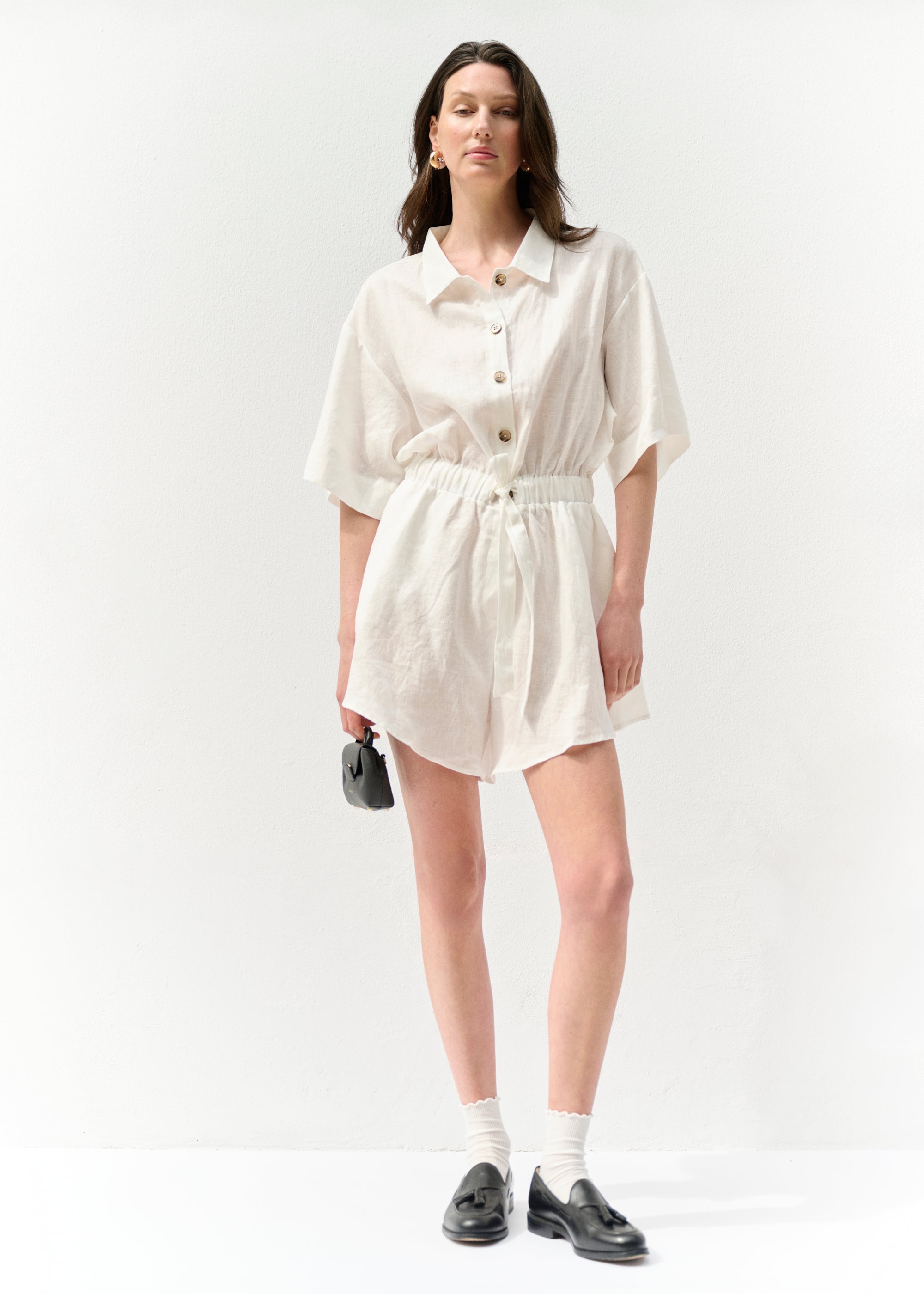 Model trägt einen weißen kurzärmeligen Leinen-Overall mit Hemdkragen und Knopfleiste vorne. Der Overall hat eine betonte Taille mit Tunnelzug und Bindeband sowie locker geschnittene Shorts. Das Model kombiniert das Outfit mit weißen Söckchen, schwarzen Loafern und goldenen Creolen, während es eine kleine schwarze Tasche in der Hand hält.