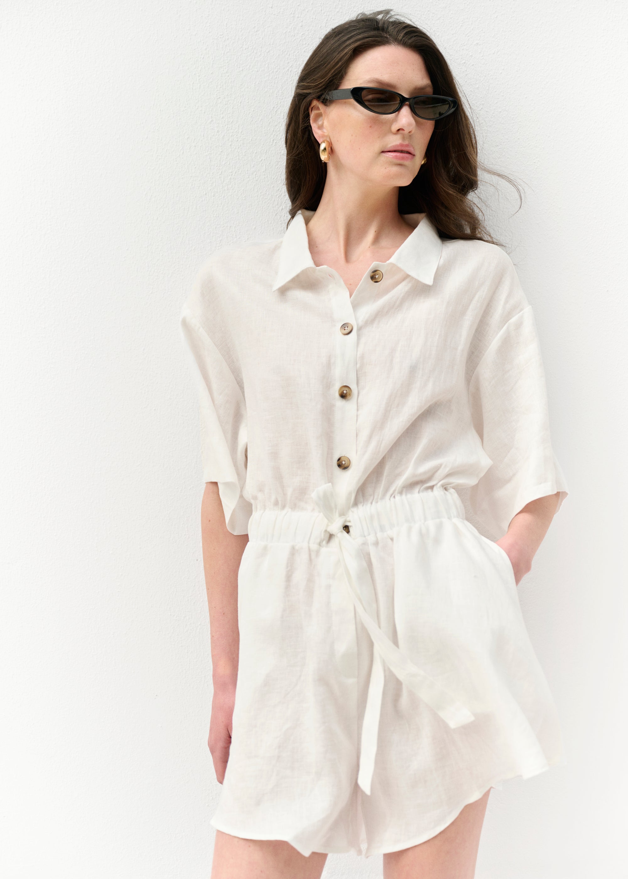 Model steht in einem weißen Leinen-Overall mit Knopfleiste und Tunnelzug-Taille. Die Kombination aus lockeren Shorts und weitem Kurzarm-Design wirkt luftig. Das Outfit wird mit schwarzer Sonnenbrille und goldenen Creolen ergänzt.