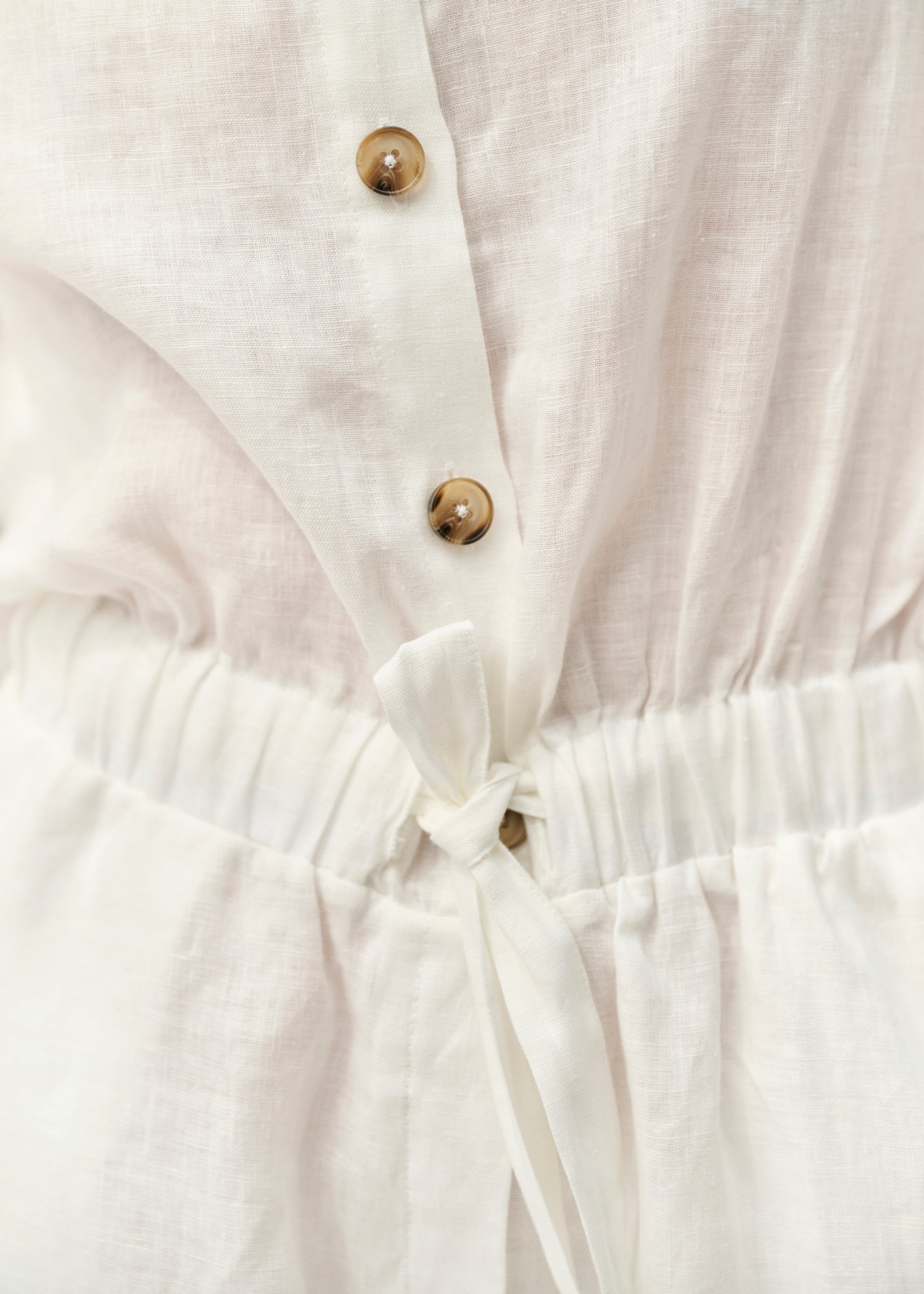 Nahaufnahme der Taille des weißen Leinen-Overalls. Zu sehen sind die Knopfleiste mit kontrastierenden Knöpfen in Hornoptik und der geraffte Bund mit weißem Bindeband.