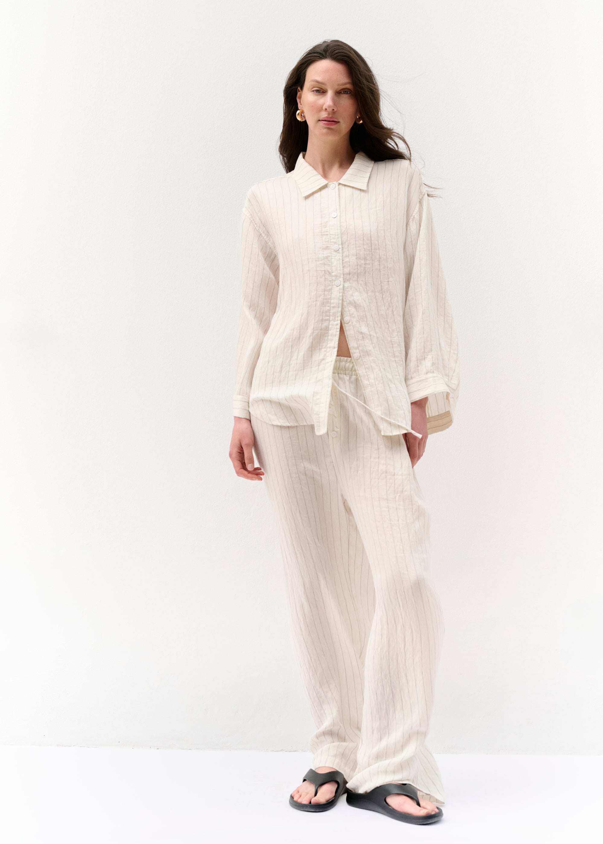 Eine Frau steht leicht frontal zur Kamera und trägt ein locker geschnittenes, cremefarbenes Zweiteiler-Set aus einem langärmligen Hemd mit Knopfleiste und einer weiten Hose. Der Stoff ist fein gestreift. Sie trägt goldene Creolen und schwarze Zehensandalen.
