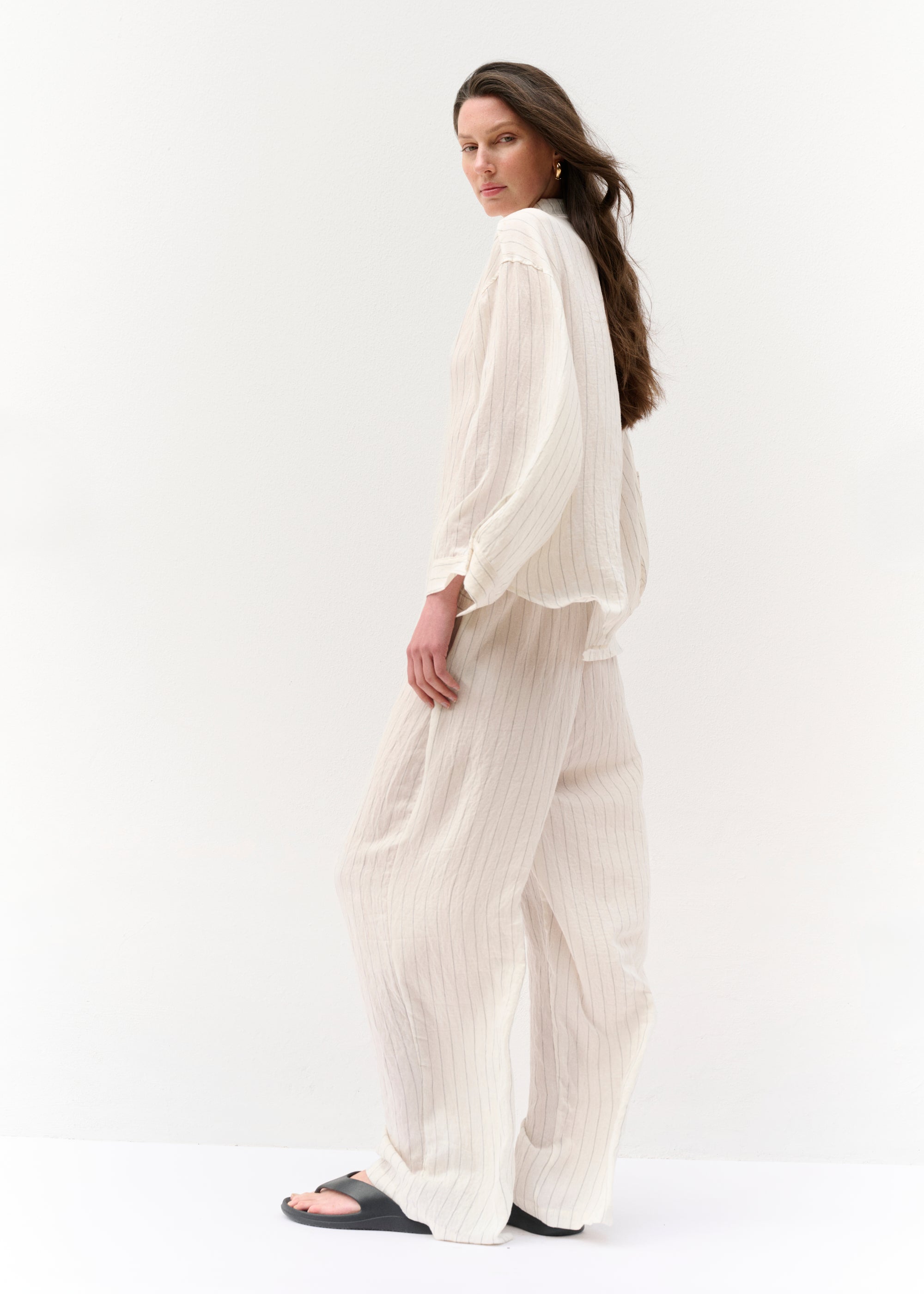 Seitlich aufgenommene Ansicht der Frau, die das gestreifte Set mit langer Hose und lockerer Hemdbluse trägt. Ihre Haare wehen locker nach hinten. Die Ärmel sind lang und leicht hochgerollt, der Look wirkt entspannt und luftig.