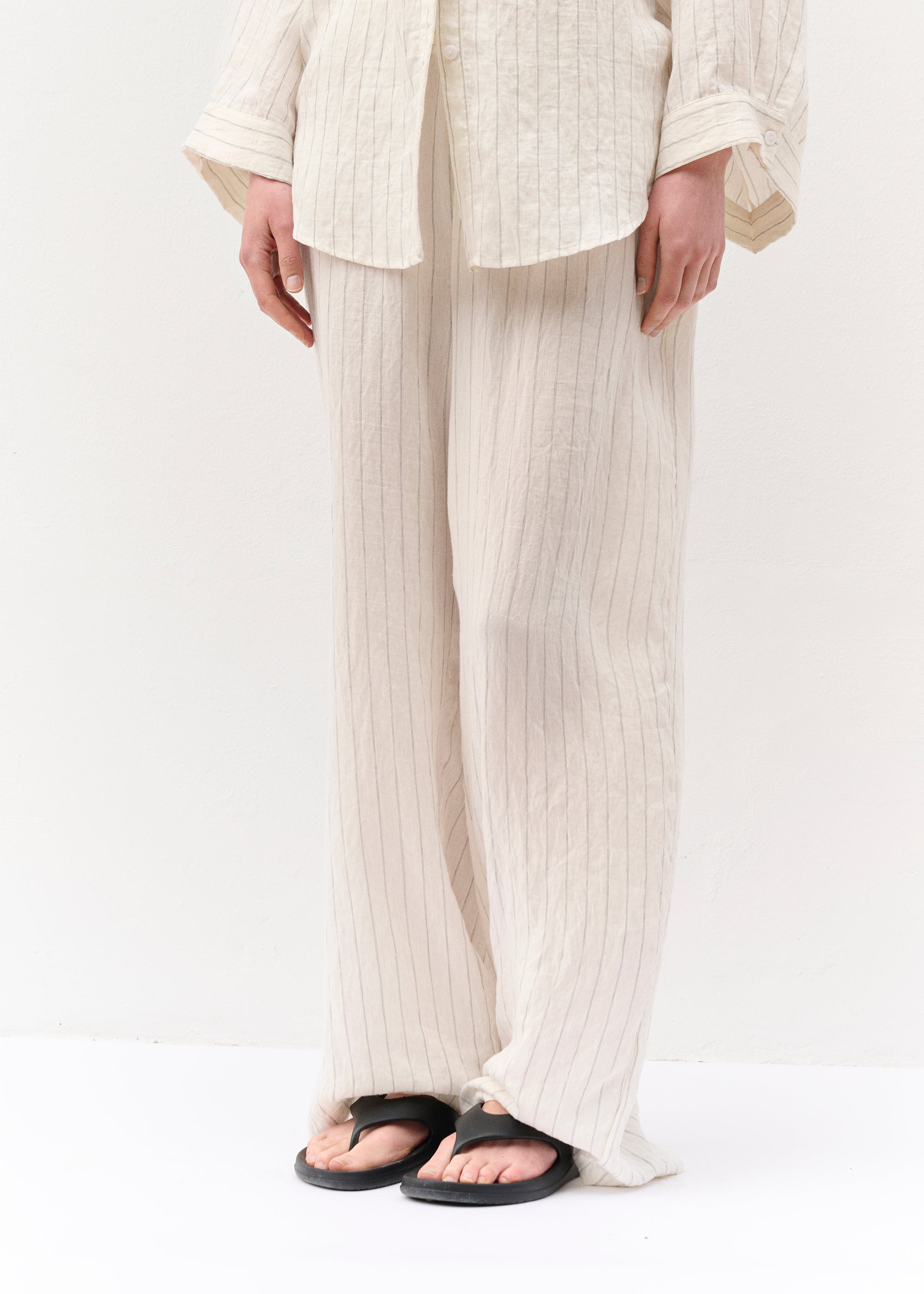 Frontalansicht des Zweiteilers in voller Länge. Die Frau trägt eine lange, gestreifte Hemdbluse mit Knöpfen und eine weite Hose aus demselben Stoff. Der Look wird durch schwarze Zehensandalen ergänzt. Die Hände hängen locker an den Seiten.