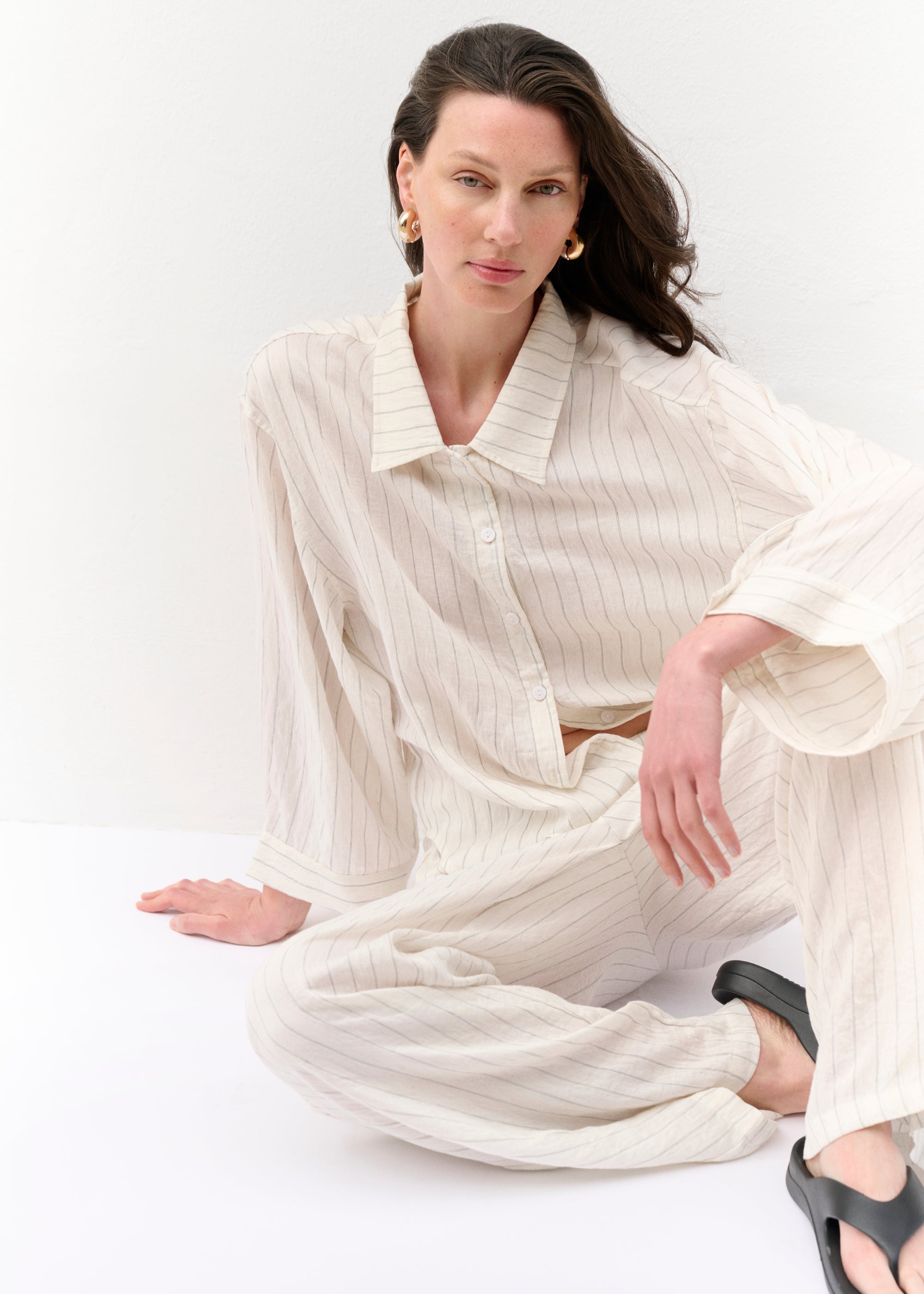 Frontalansicht des Zweiteilers in voller Länge. Die Frau trägt eine lange, gestreifte Hemdbluse mit Knöpfen und eine weite Hose aus demselben Stoff. Der Look wird durch schwarze Zehensandalen ergänzt. Die Hände hängen locker an den Seiten.