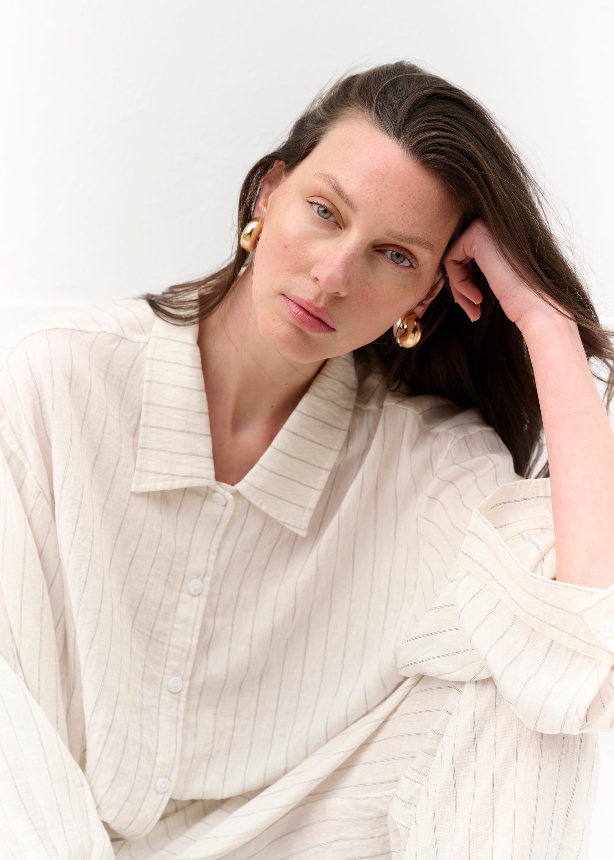 Eine Frau sitzt entspannt auf dem Boden vor einer weißen Wand. Sie trägt ein cremefarbenes, gestreiftes Set aus locker fallender Hemdbluse mit Knopfleiste und weiter Hose. Die langen Ärmel sind ausgestellt, der Stoff leicht transparent. Sie trägt schwarze Slides und goldene Creolen. Ihr Blick ist ruhig und direkt zur Kamera gerichtet.