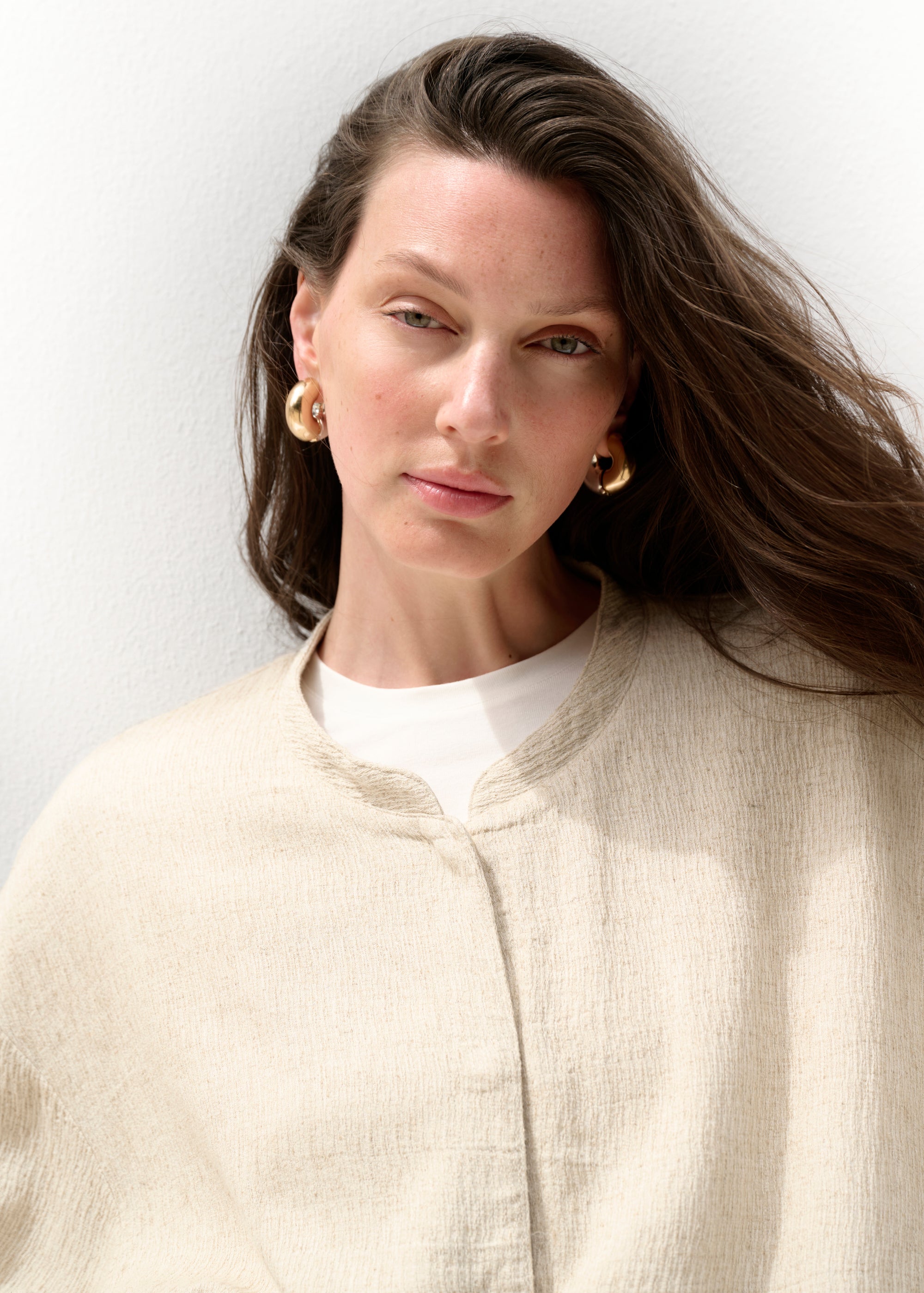 Nahaufnahme des Gesichts der Frau. Sie trägt goldene Creolen, ihre Haare wehen leicht im Wind. Unter der beigen Jacke ist ein weißer T-Shirt-Ausschnitt zu erkennen. Ihr Blick ist ruhig und direkt in die Kamera gerichtet.
