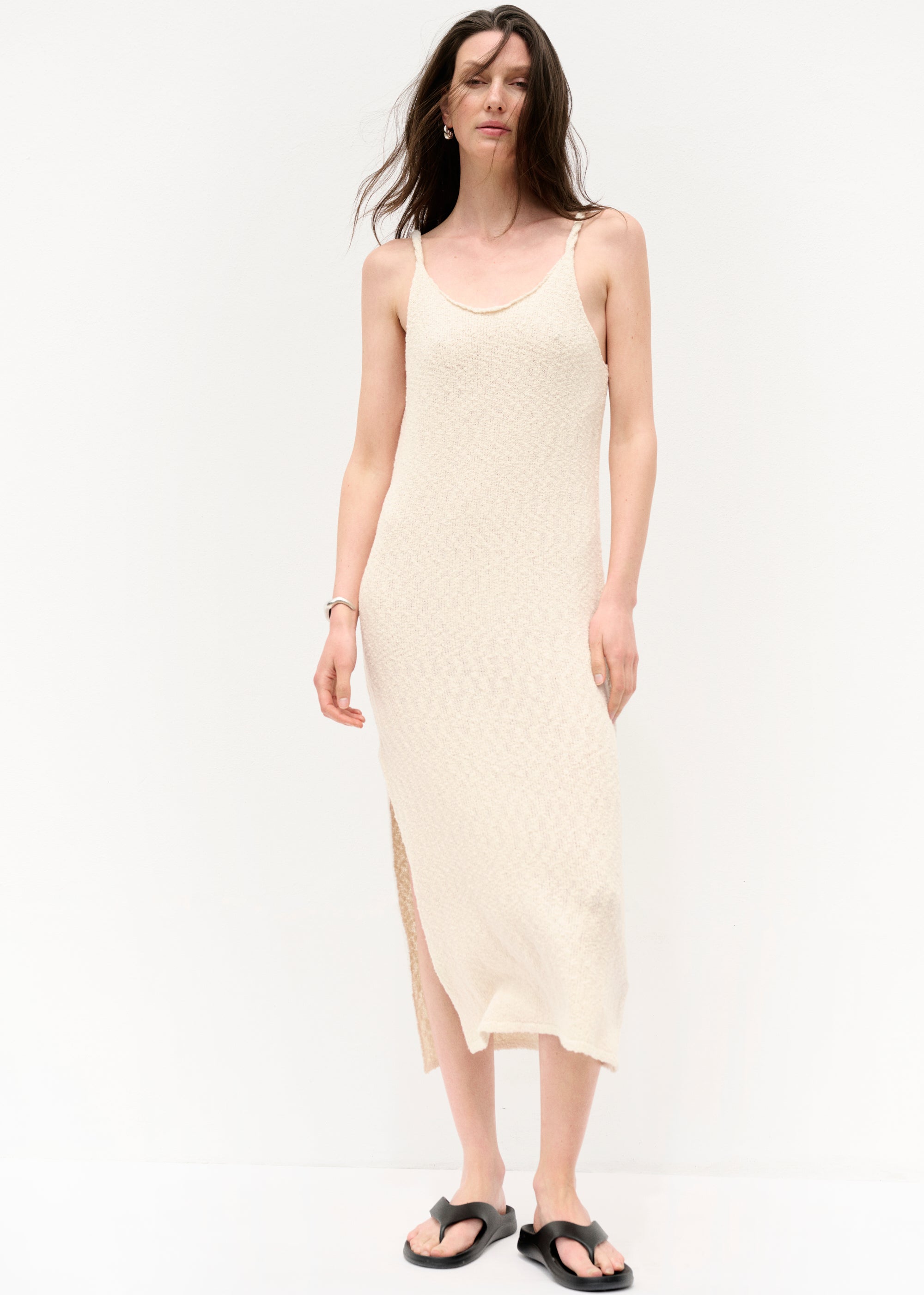 Frau in einem ärmellosen, cremefarbenen Strickkleid mit Spaghettiträgern und seitlichem Schlitz, barfuß in schwarzen Sandalen, vor hellem Hintergrund stehend, mit offenem Haar und neutralem Gesichtsausdruck.