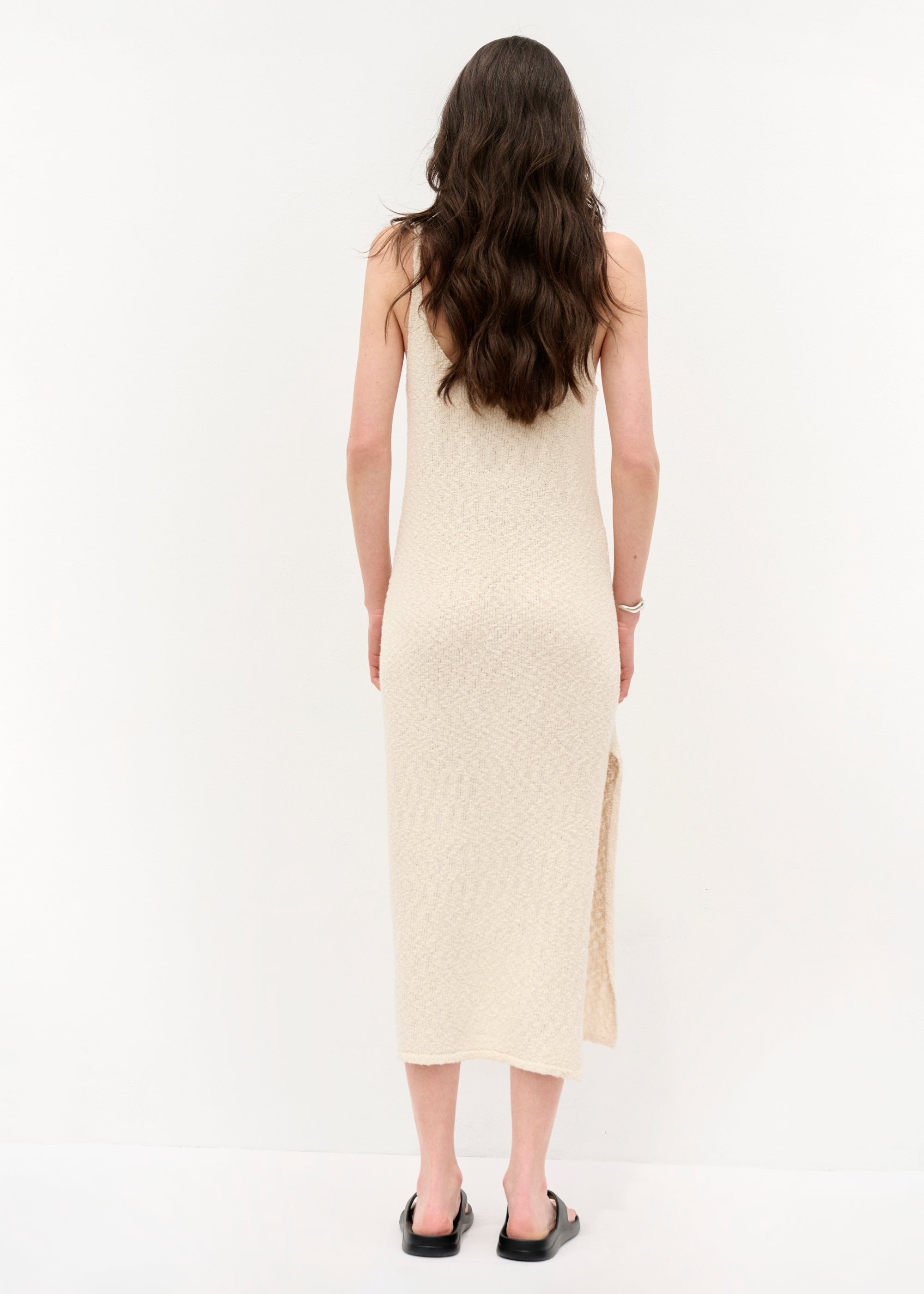 Frau von hinten in einem ärmellosen, cremefarbenen Strickkleid mit Spaghettiträgern und seitlichem Schlitz, barfuß in schwarzen Sandalen, vor hellem Hintergrund stehend, mit offenem Haar .