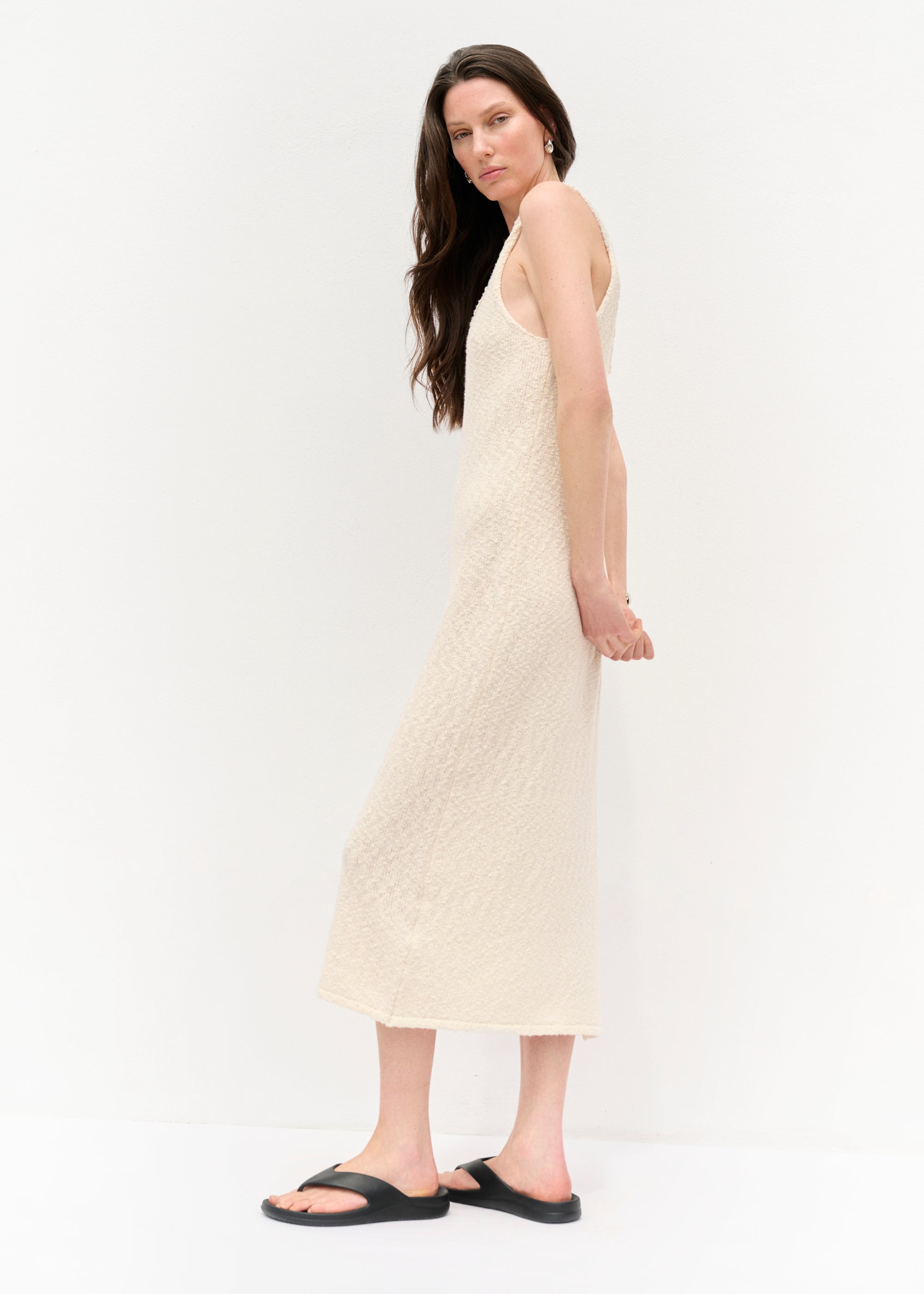 Frau in einem ärmellosen, cremefarbenen Strickkleid mit Spaghettiträgern und seitlichem Schlitz, barfuß in schwarzen Sandalen, vor hellem Hintergrund stehend, mit offenem Haar und neutralem Gesichtsausdruck.