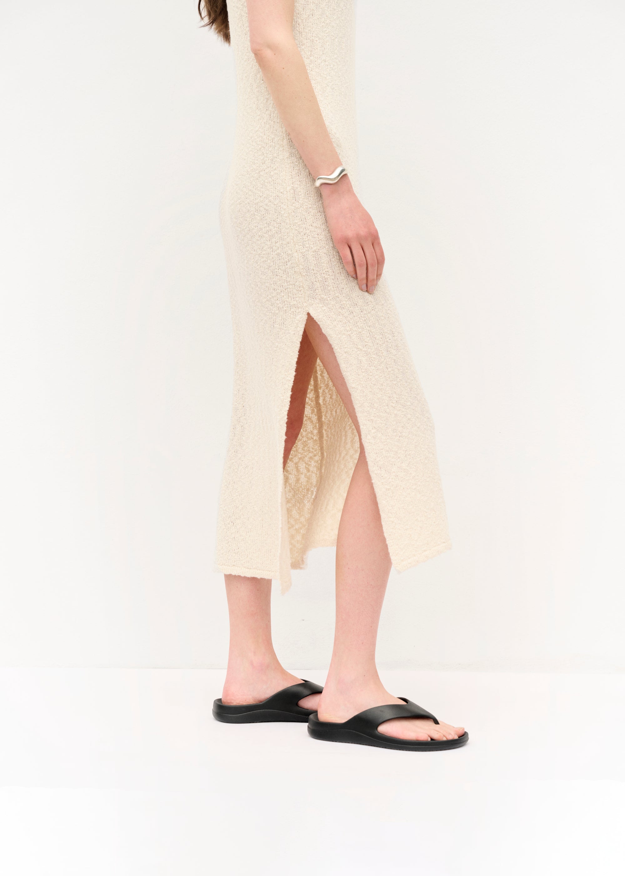 Frau in einem ärmellosen, cremefarbenen Strickkleid mit Spaghettiträgern und seitlichem Schlitz, barfuß in schwarzen Sandalen, vor hellem Hintergrund stehend, mit offenem Haar.