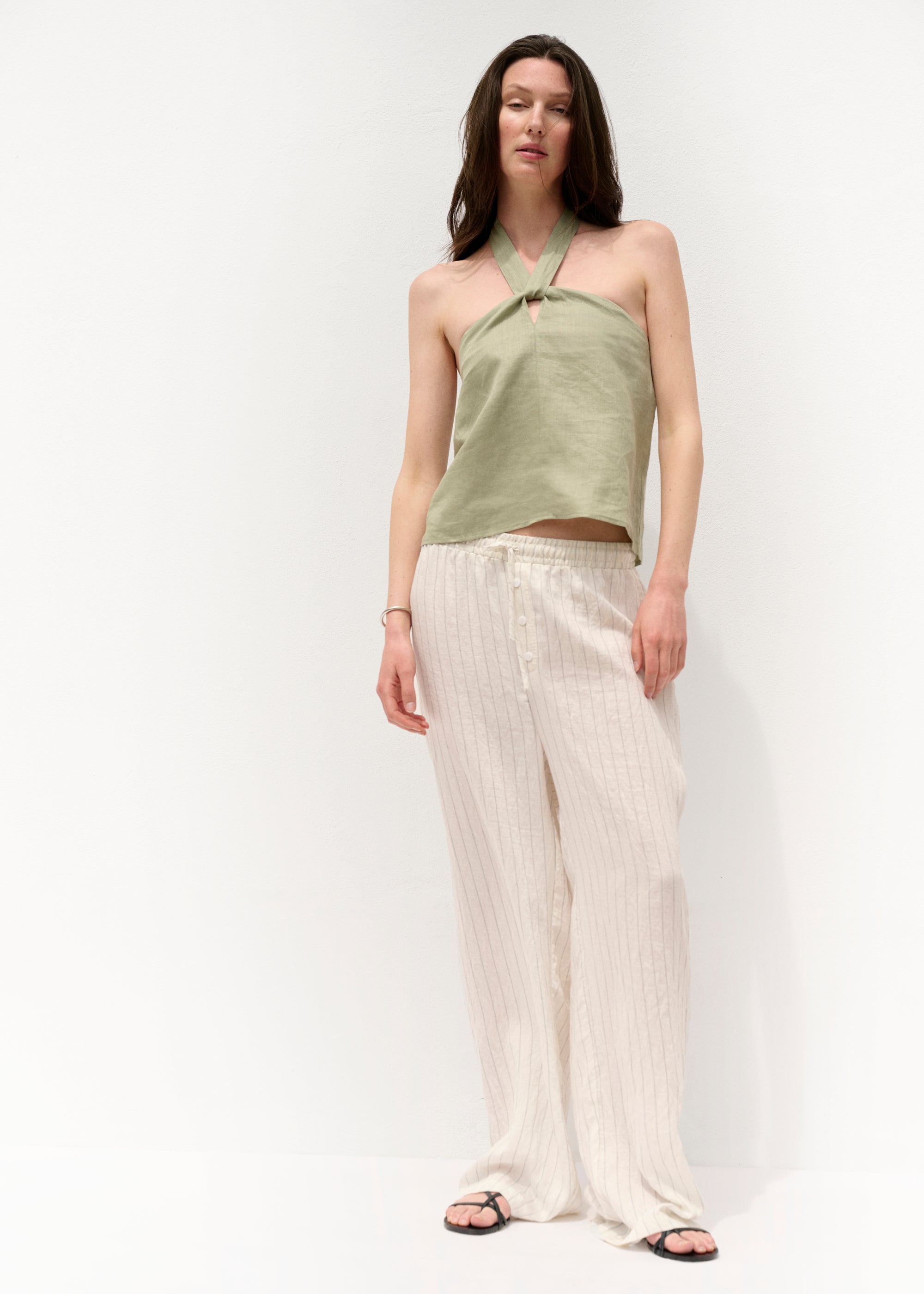 Vollansicht von vorne: Die Frau trägt das olivegrüne Neckholder-Top zusammen mit einer weiten, cremefarbenen Hose mit feinen vertikalen Streifen. Ihr Stand ist entspannt, der Hintergrund ist neutral.