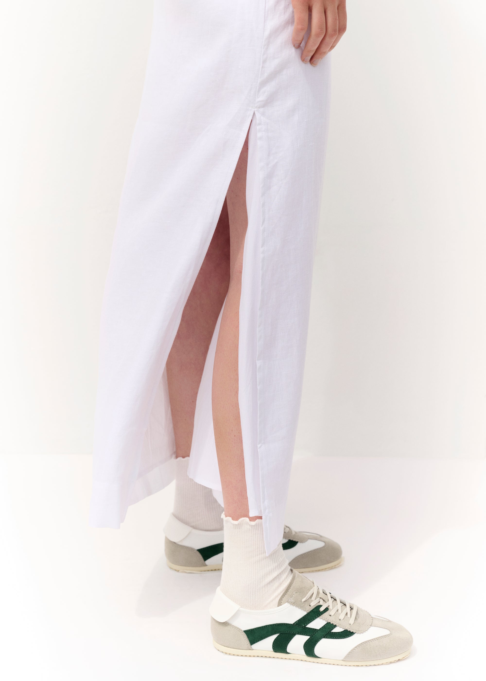 Nahaufnahme des weißen Leinenrocks mit seitlichem Schlitz, der bis zum Knie reicht. Sichtbar sind außerdem die beigefarbenen Retro-Sneaker mit grünen Streifendetails sowie gerippte cremefarbene Socken mit gewelltem Abschluss.