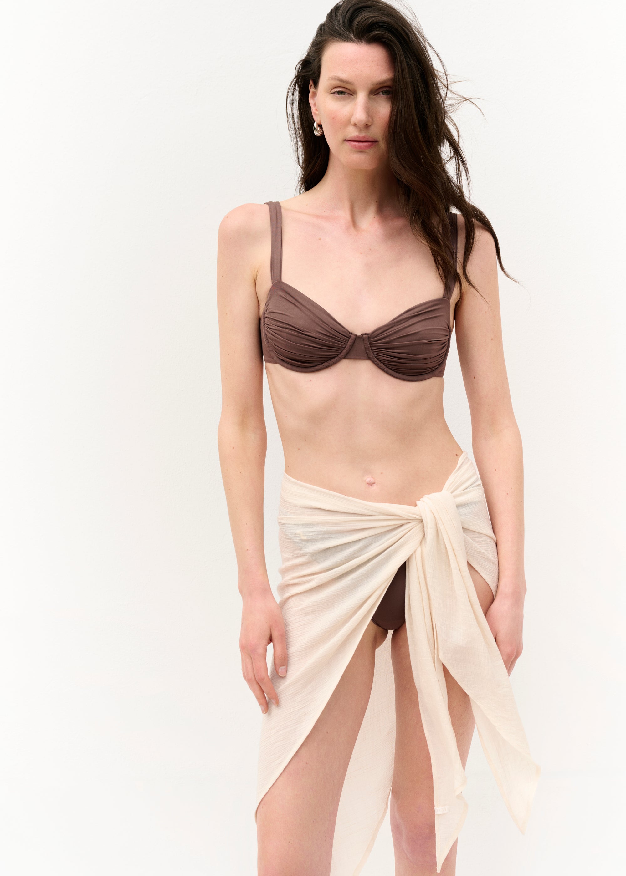 Eine Frau mit langen dunklen Haaren trägt einen kurzen, beigefarbenen Pareo aus leichtem, transparentem Stoff, der seitlich geknotet ist. Darunter ist ein brauner Bikini zu sehen. Die Fotos zeigen sie frontal, von hinten und aus der Nähe vor neutralem, weißem Hintergrund.