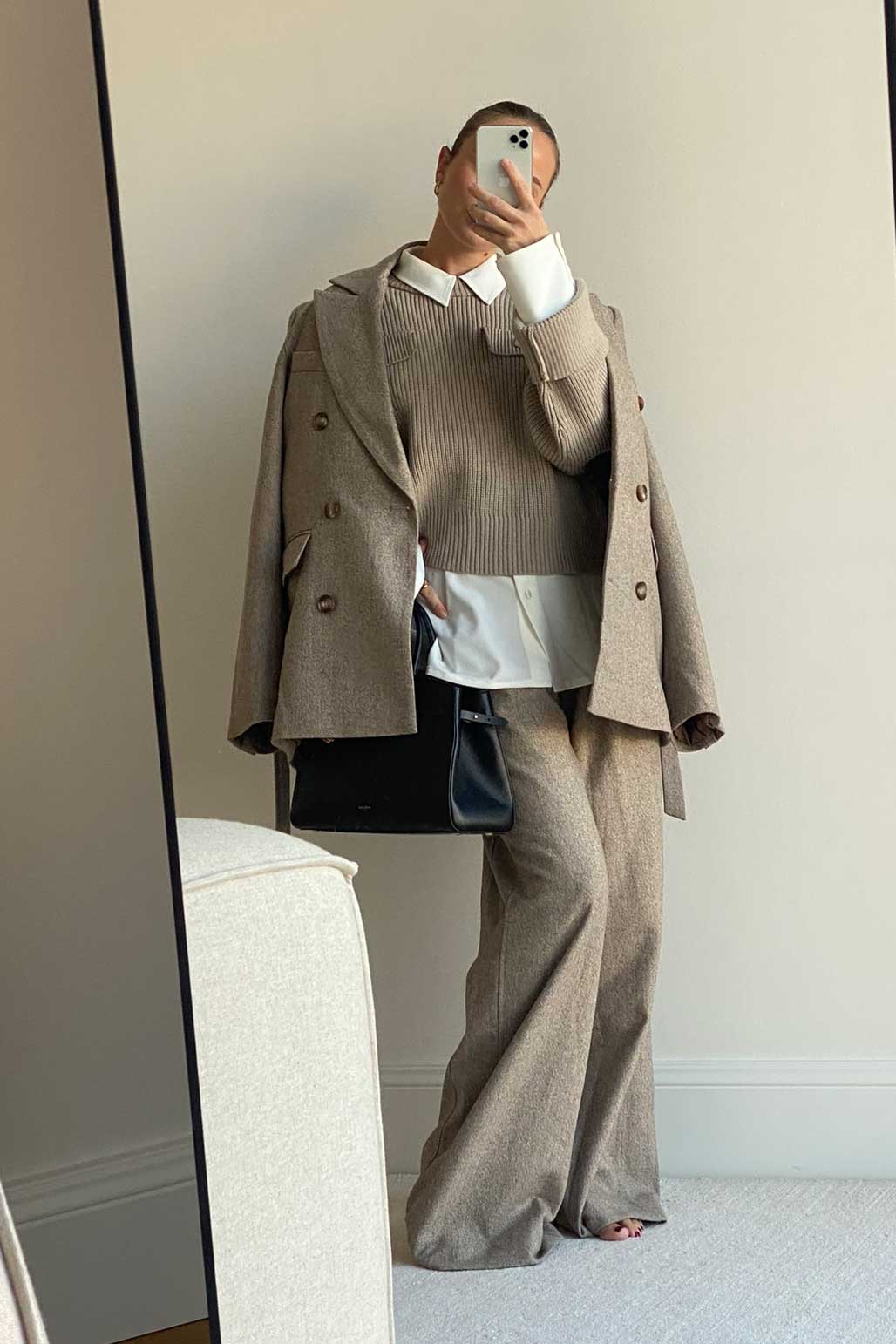 Frau steht in einem eleganten Business-Look vor einem Spiegel. Sie trägt eine beige Wollhose mit weitem Bein, eine weiße Hemdbluse unter einem beigefarbenen, gerippten Strickpullover mit Brusttaschen und hat einen passenden Wollblazer locker über die Schultern gelegt. In der Hand hält sie eine schwarze Tasche. Das Outfit wirkt herbstlich und stilvoll.