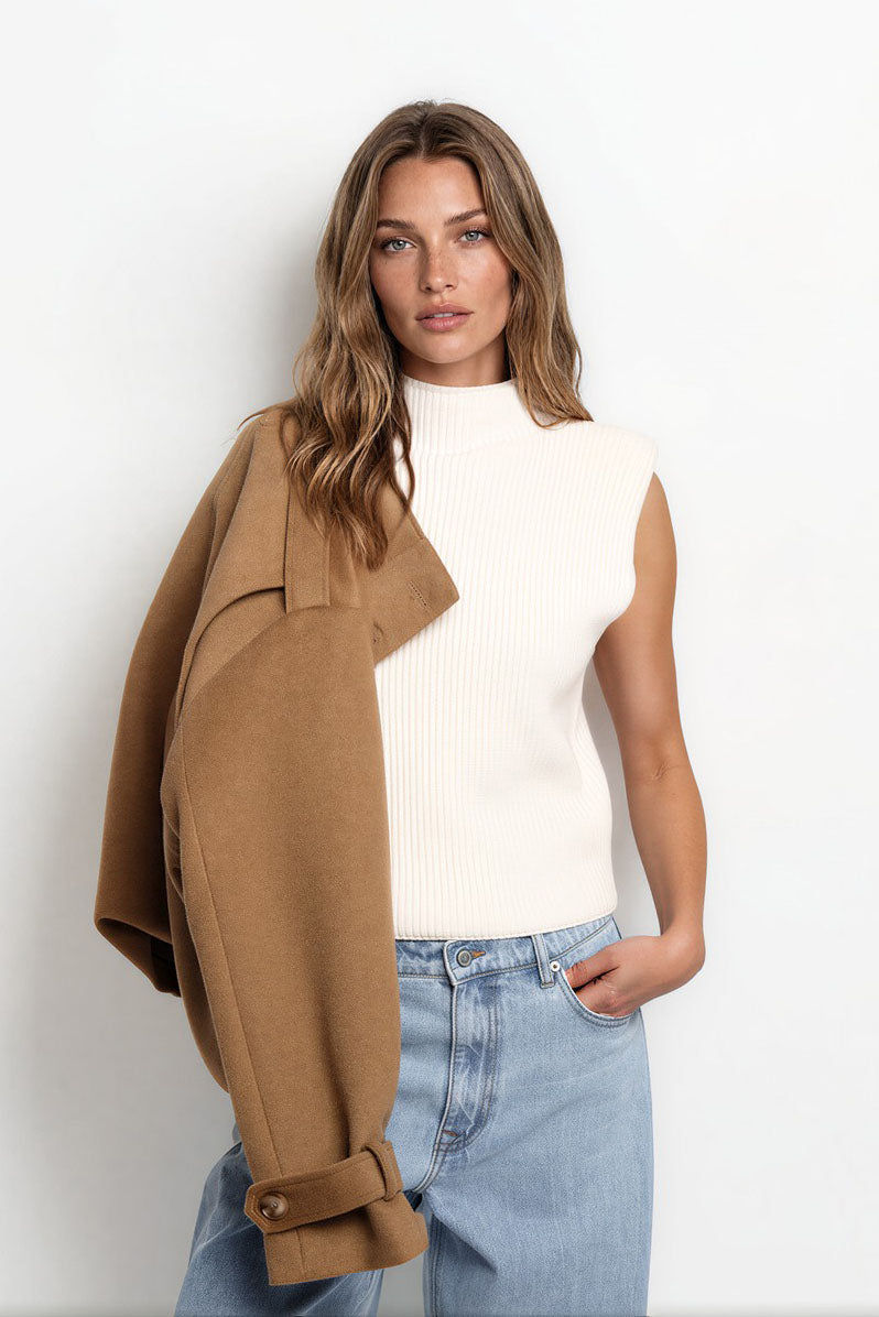 Frau trägt ein cremeweißes ärmelloses Stricktop mit Stehkragen zu einer hellblauen Jeans und hält einen camel-farbenen Mantel über der Schulter.