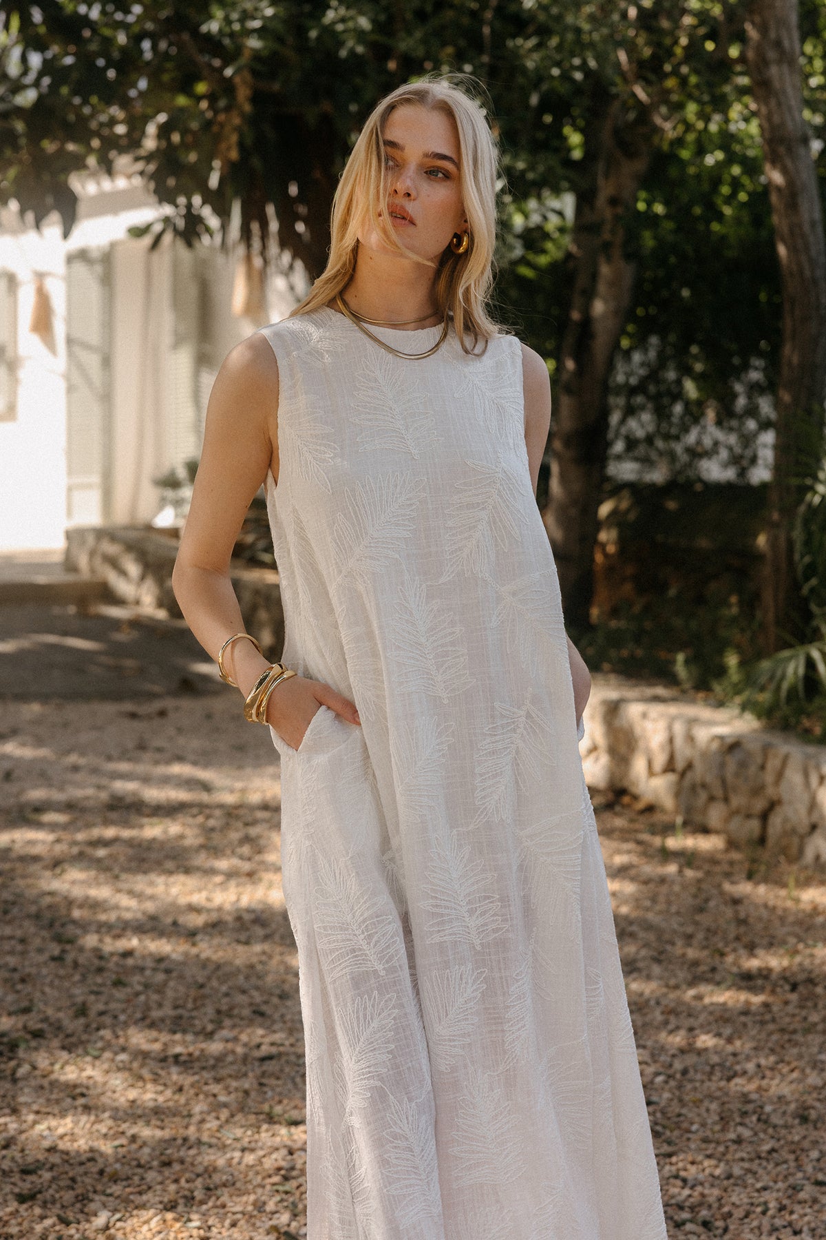 Model in langem weißen Maxikleid mit feiner Blattstickerei und seitlichen Taschen auf einem sonnigen Gartenweg.