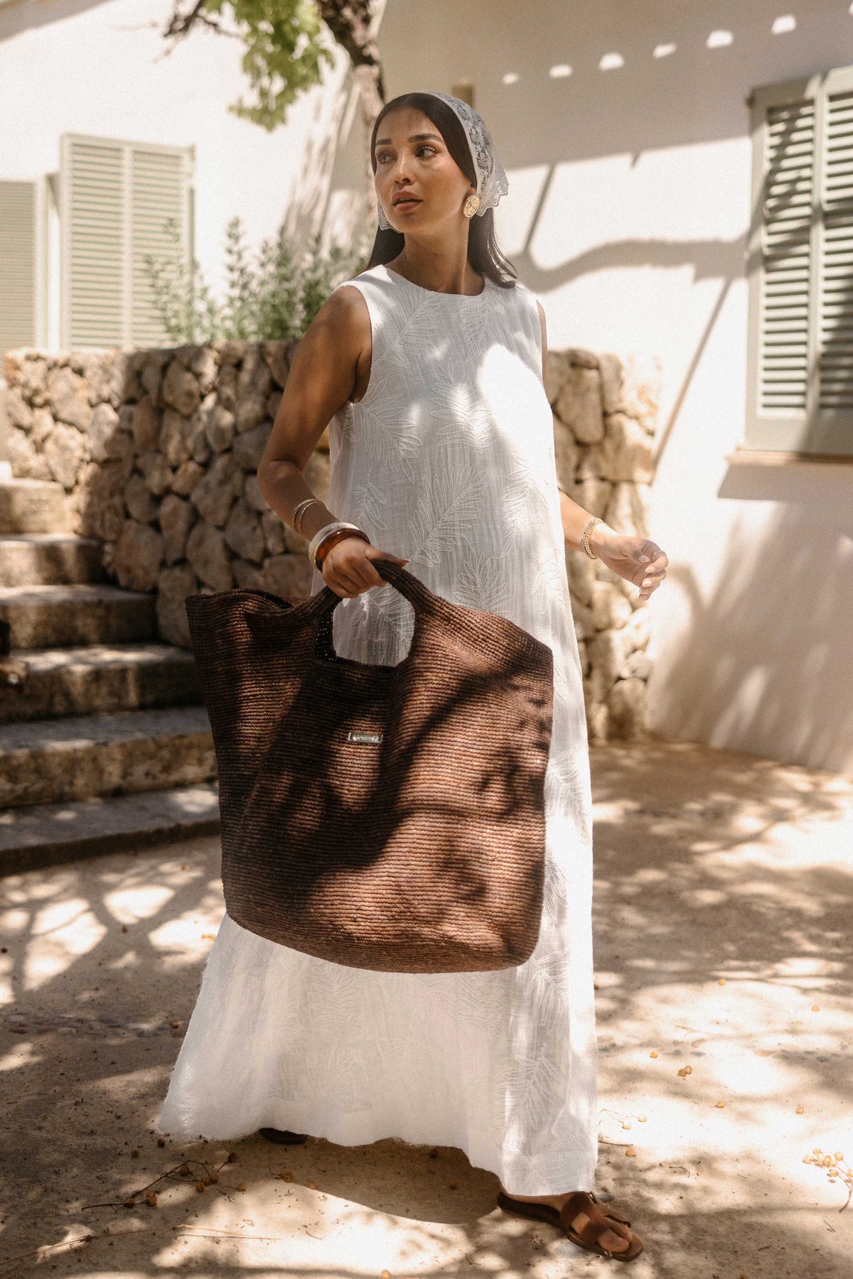 Frau in einem weißen, ärmellosen Minikleid mit eingewebtem Blattmuster steht im Schatten mediterraner Bäume vor einem Landhaus. Sie trägt goldene Ohrringe, eine kleine braune Schultertasche und blickt zur Seite. Das warme Sonnenlicht erzeugt ein natürliches Schattenspiel auf Kleid und Umgebung.