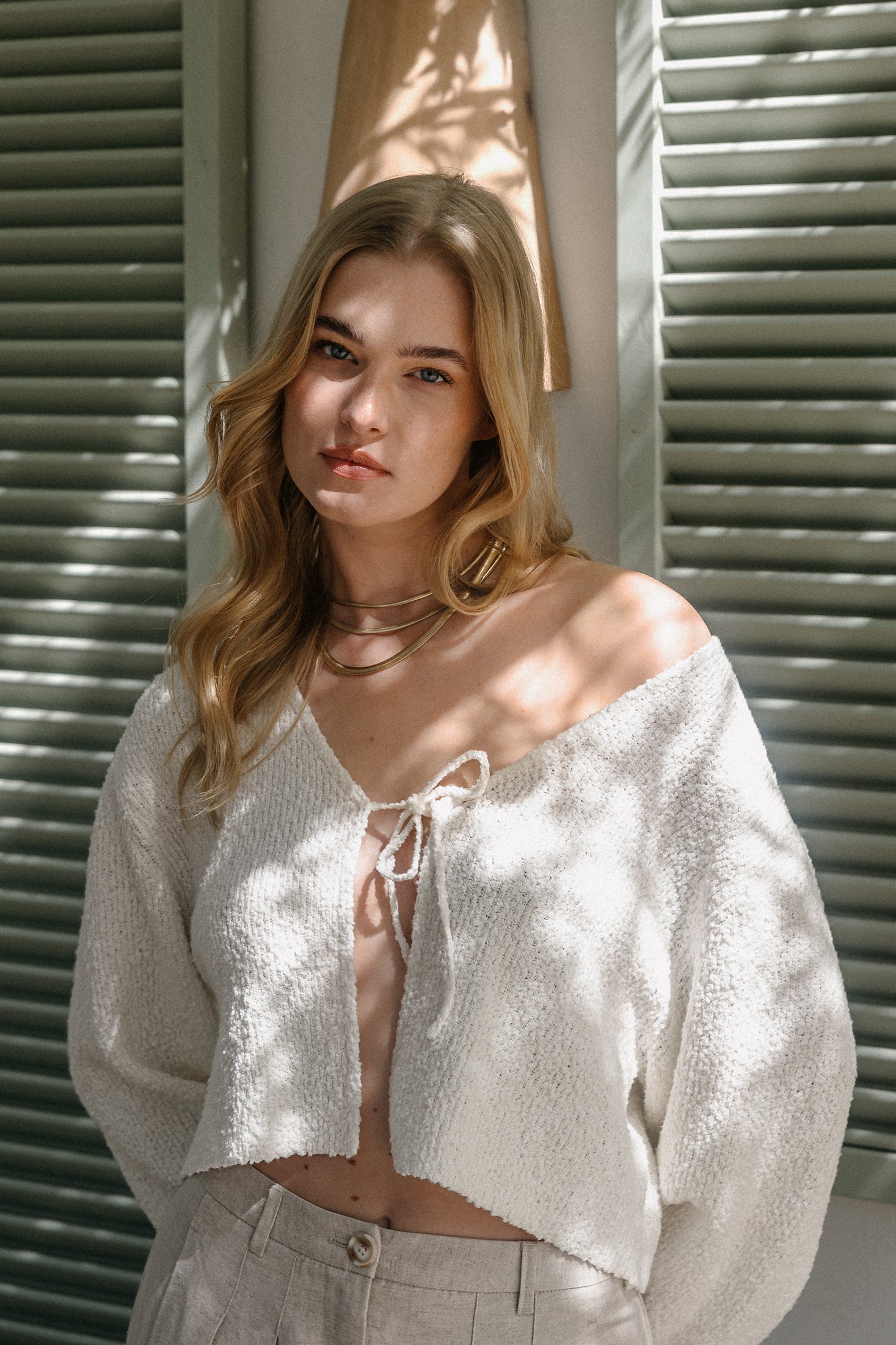 Model trägt einen weißen, strukturierten Strick-Cardigan mit Bindeband und V-Ausschnitt, kombiniert mit einer beigen Hose, im warmen Licht vor Fensterläden