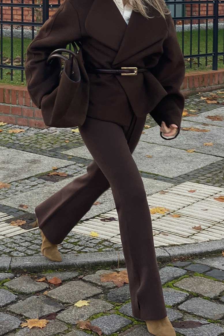 Frau im Gehen, gekleidet in ein monochromes Ensemble aus schokoladenbraunem Wollblazer mit Gürtel und farblich abgestimmter, weit geschnittener Hose mit Schlitz am Saum; dazu beigefarbene Stiefeletten und eine passende braune Handtasche. Aufgenommen auf herbstlichem Straßenpflaster mit Laub.