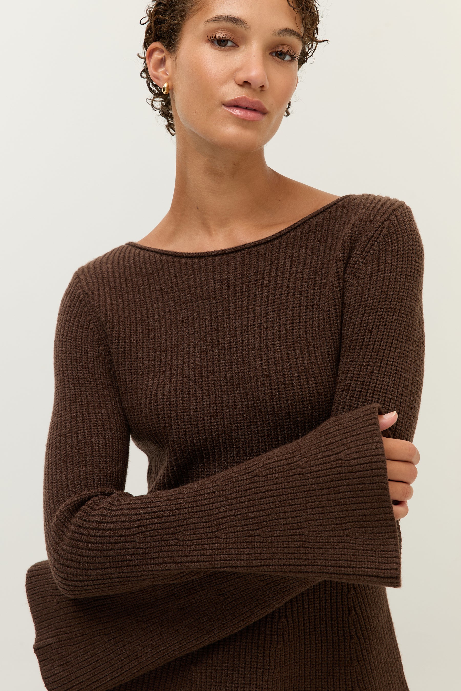 Frau trägt ein lockeres, zweiteiliges Set aus strukturiertem Bouclé-Stoff in dunklem Schokobraun. Das Set besteht aus einem langärmeligen, weiten Pullover mit Rundhalsausschnitt und einer passenden, weiten Hose mit geradem Bein. Sie kombiniert das Outfit mit goldenen Creolen und schwarzen Loafern. Der Look wirkt gemütlich und entspannt.