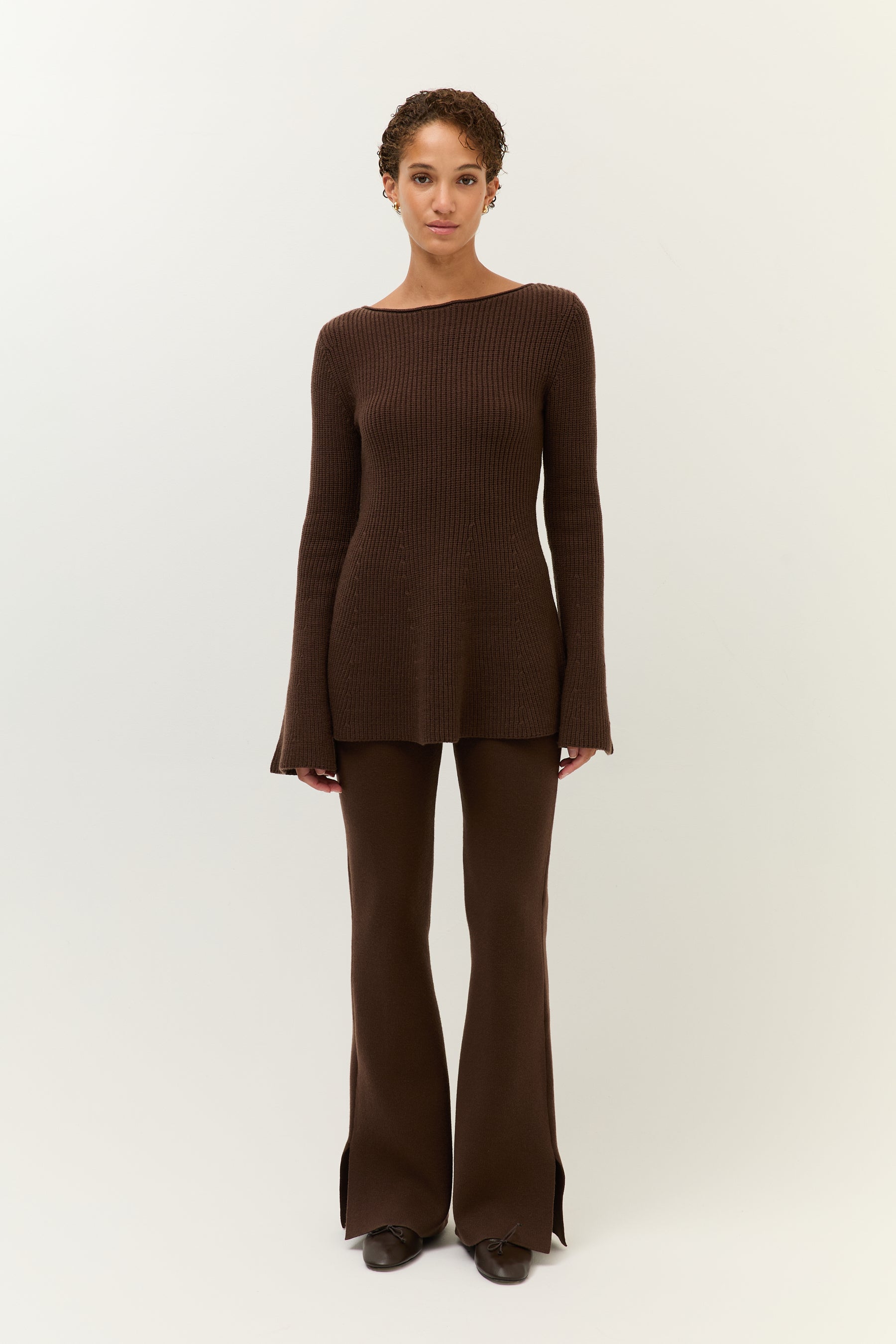 Eine Frau steht frontal vor hellem Hintergrund und trägt ein braunes, geripptes Strickoberteil mit ausgestellten Ärmeln und eine passende Schlaghose mit Schlitz am Saum.