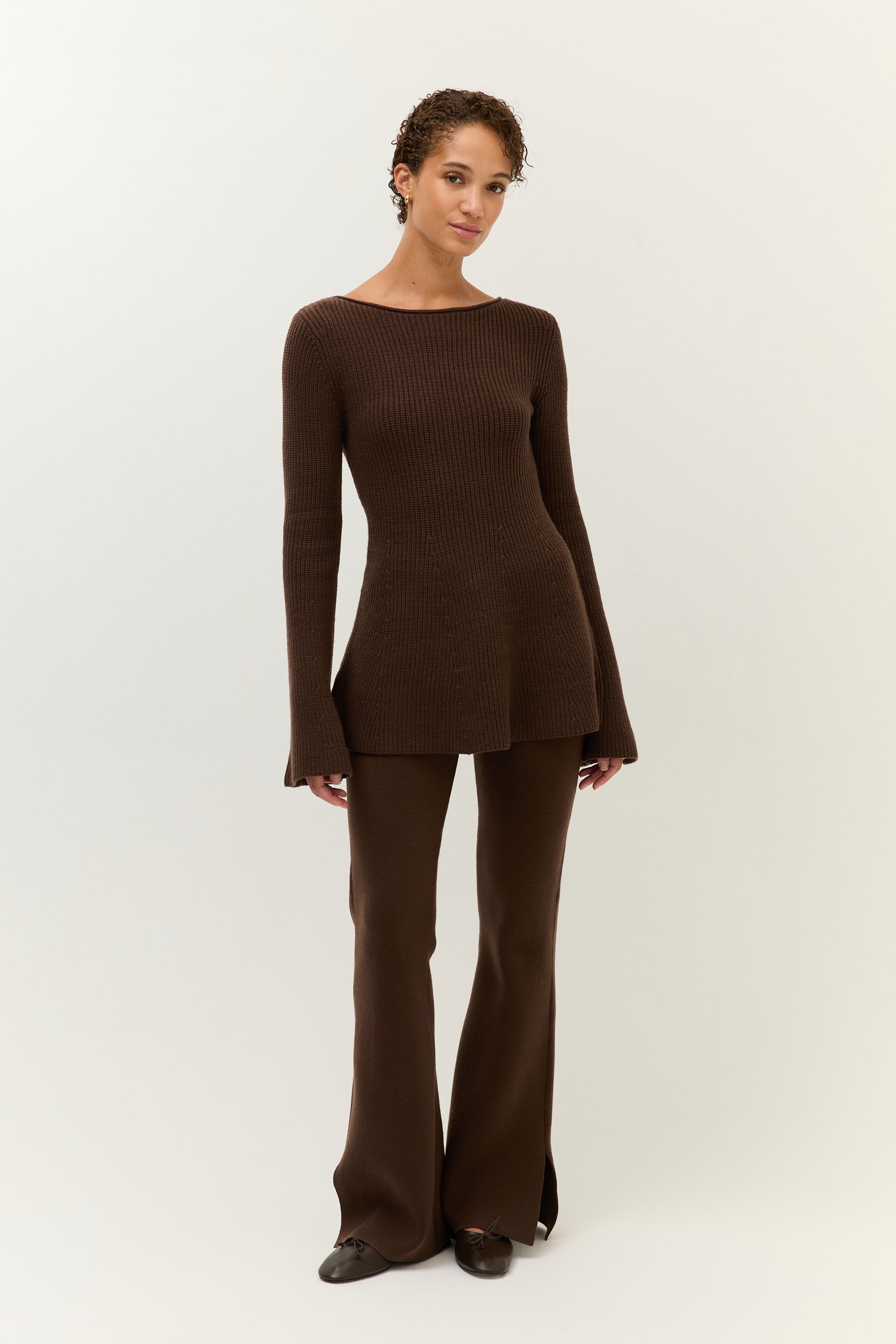 Frau trägt ein figurbetontes, zweiteiliges Set aus geripptem Strick in dunklem Braun. Das Oberteil hat einen Boot-Ausschnitt, ausgestellte Ärmel und ein leicht ausgestelltes Schößchen. Die Hose ist ebenfalls aus Strick und hat ein weites, ausgestelltes Bein. Dazu trägt sie schlichte Ballerinas und goldene Ohrringe. Der Look ist elegant und komfortabel zugleich.