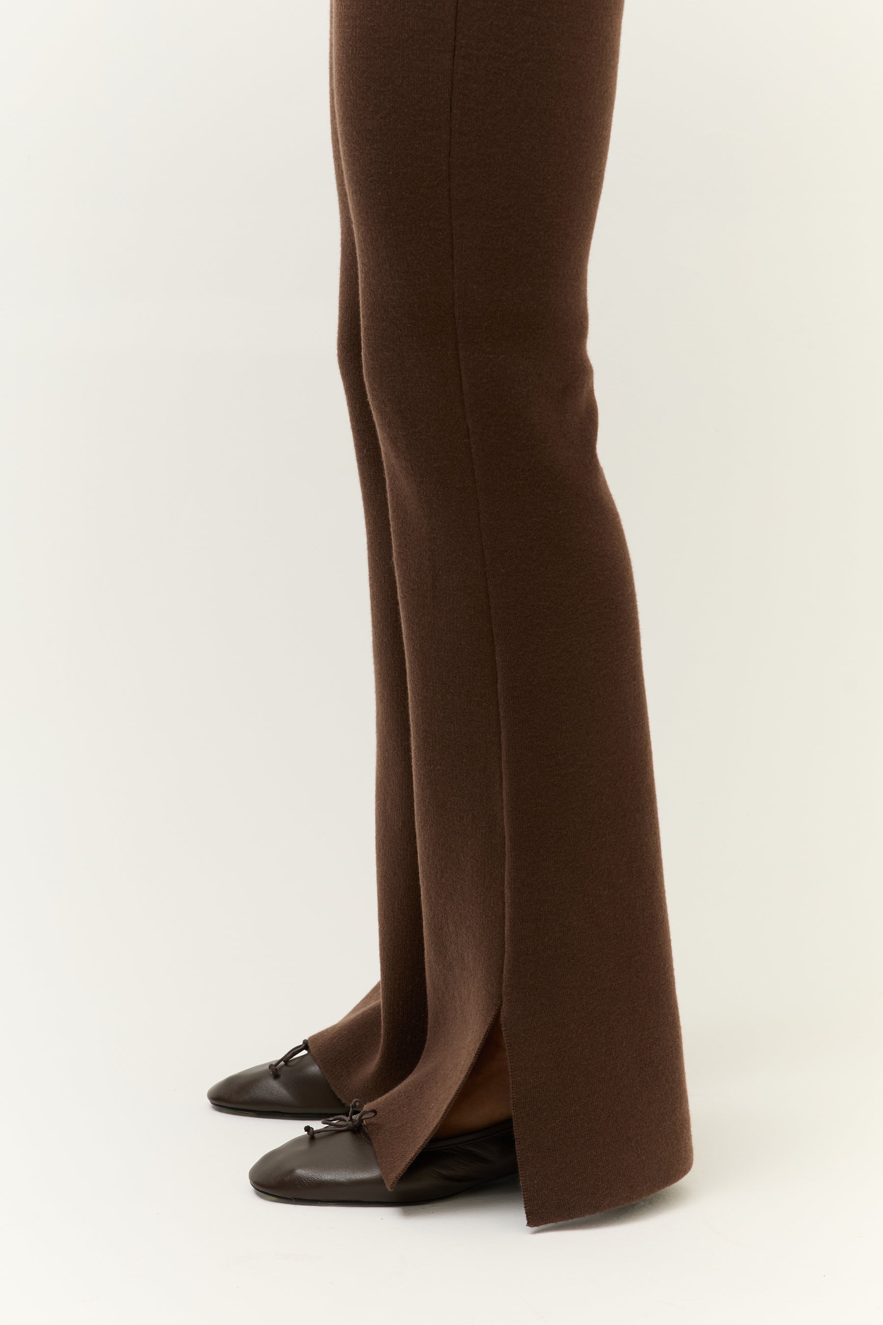 Nahaufnahme der unteren Beinpartie und Schuhe: Die braune Hose endet in einem frontalen Schlitz, darunter sind dunkelbraune Ballerinas mit Schleife sichtbar.