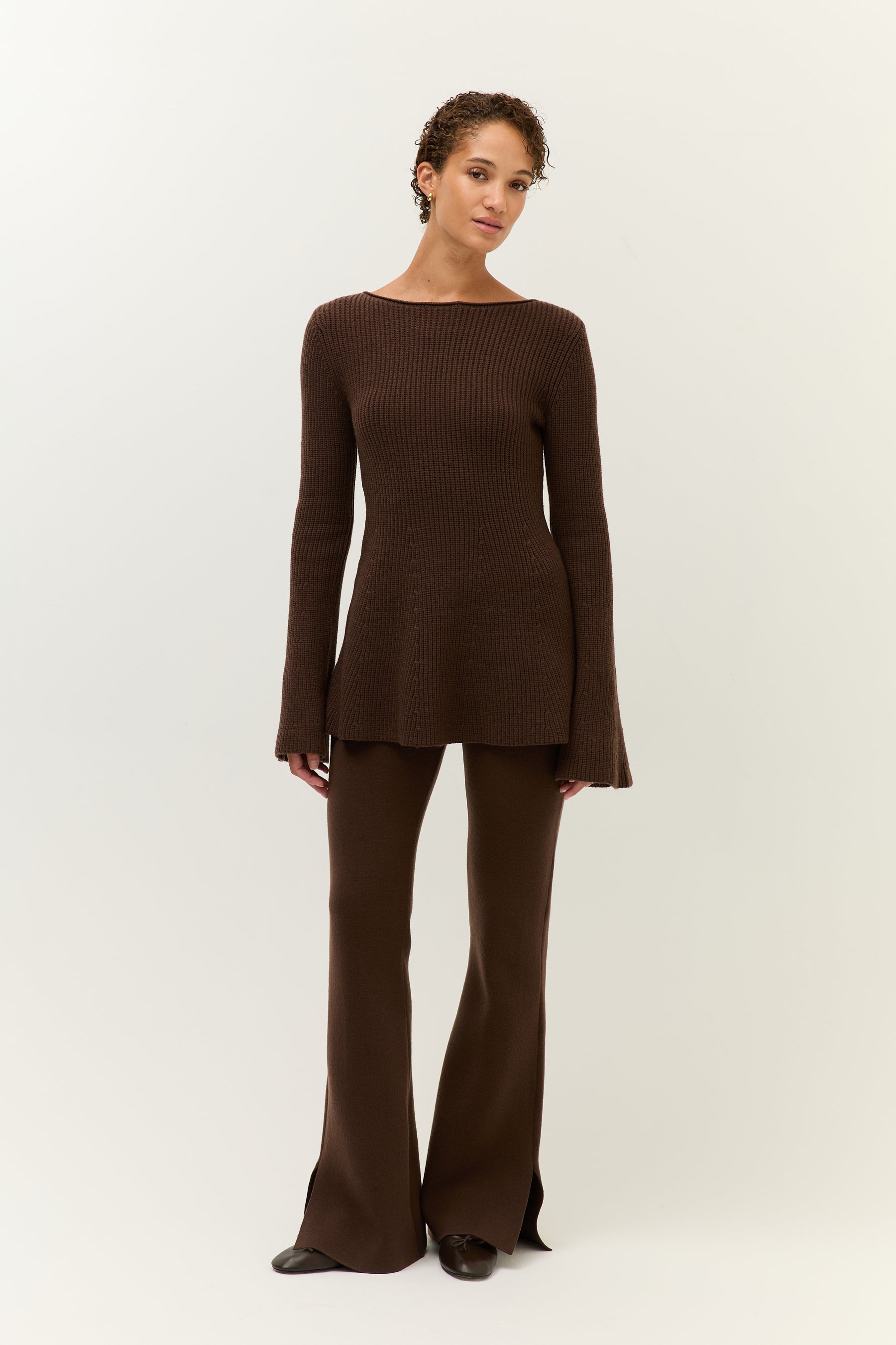 Die Frau steht leicht seitlich gedreht und blickt in die Kamera. Das braune Strickoberteil fällt locker über die Hüften, die Hose liegt figurbetont an.
