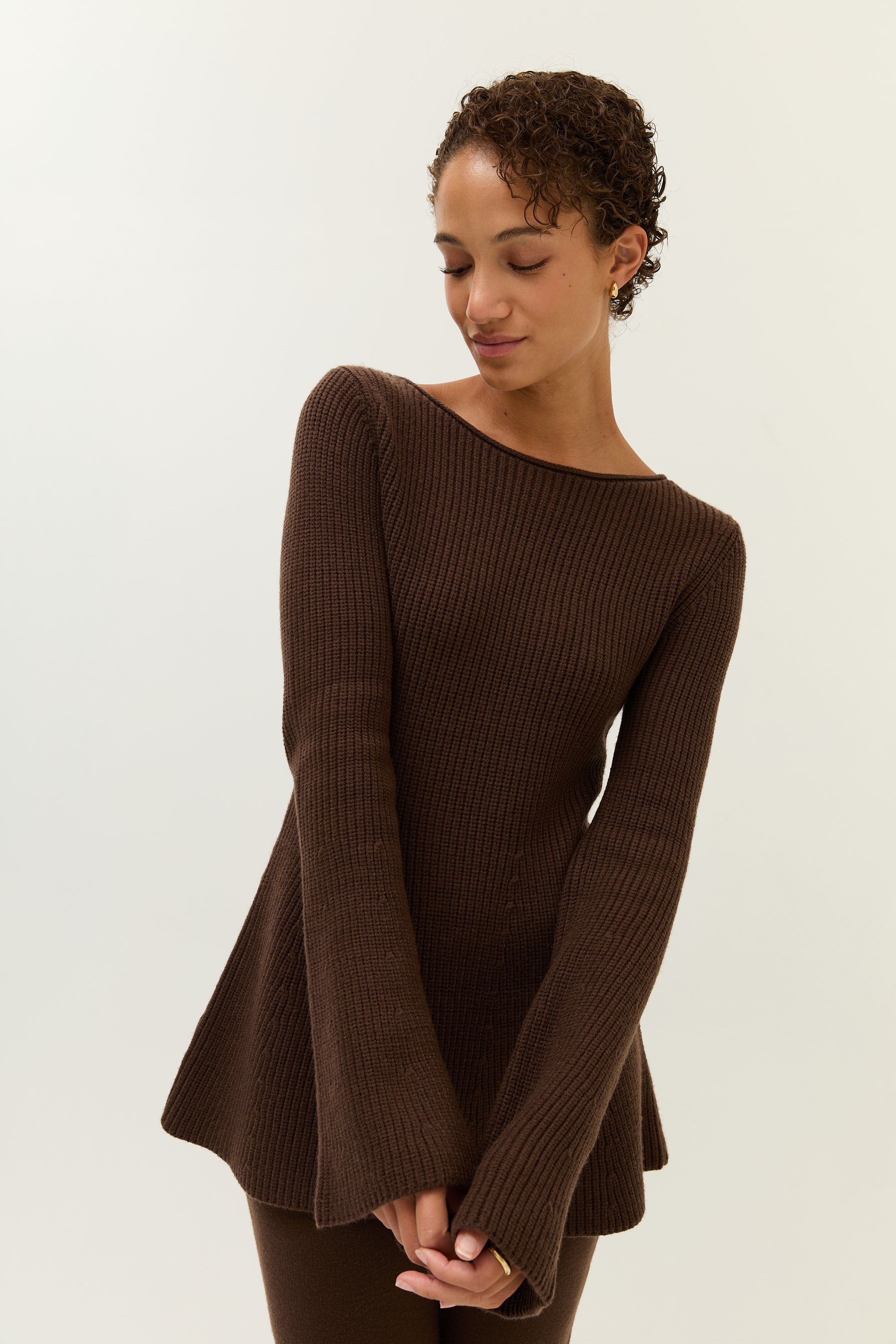 Frau trägt ein lockeres, zweiteiliges Set aus strukturiertem Bouclé-Stoff in dunklem Schokobraun. Das Set besteht aus einem langärmeligen, weiten Pullover mit Rundhalsausschnitt und einer passenden, weiten Hose mit geradem Bein. Sie kombiniert das Outfit mit goldenen Creolen und schwarzen Loafern. Der Look wirkt gemütlich und entspannt.