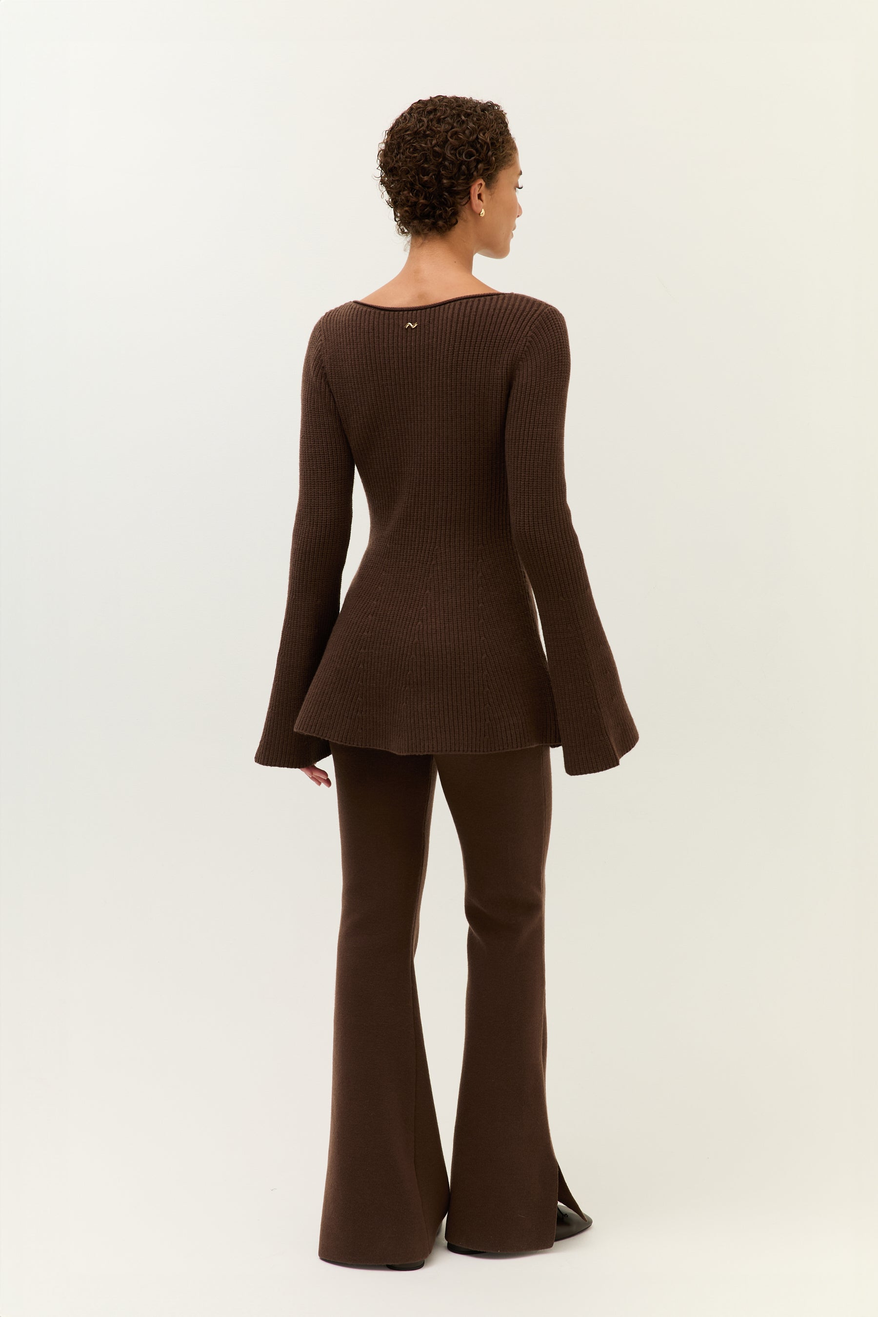 Rückansicht des gesamten Looks. Das Strickoberteil ist tailliert geschnitten und schwingt leicht aus, die Hose verläuft körpernah bis zum Saum.