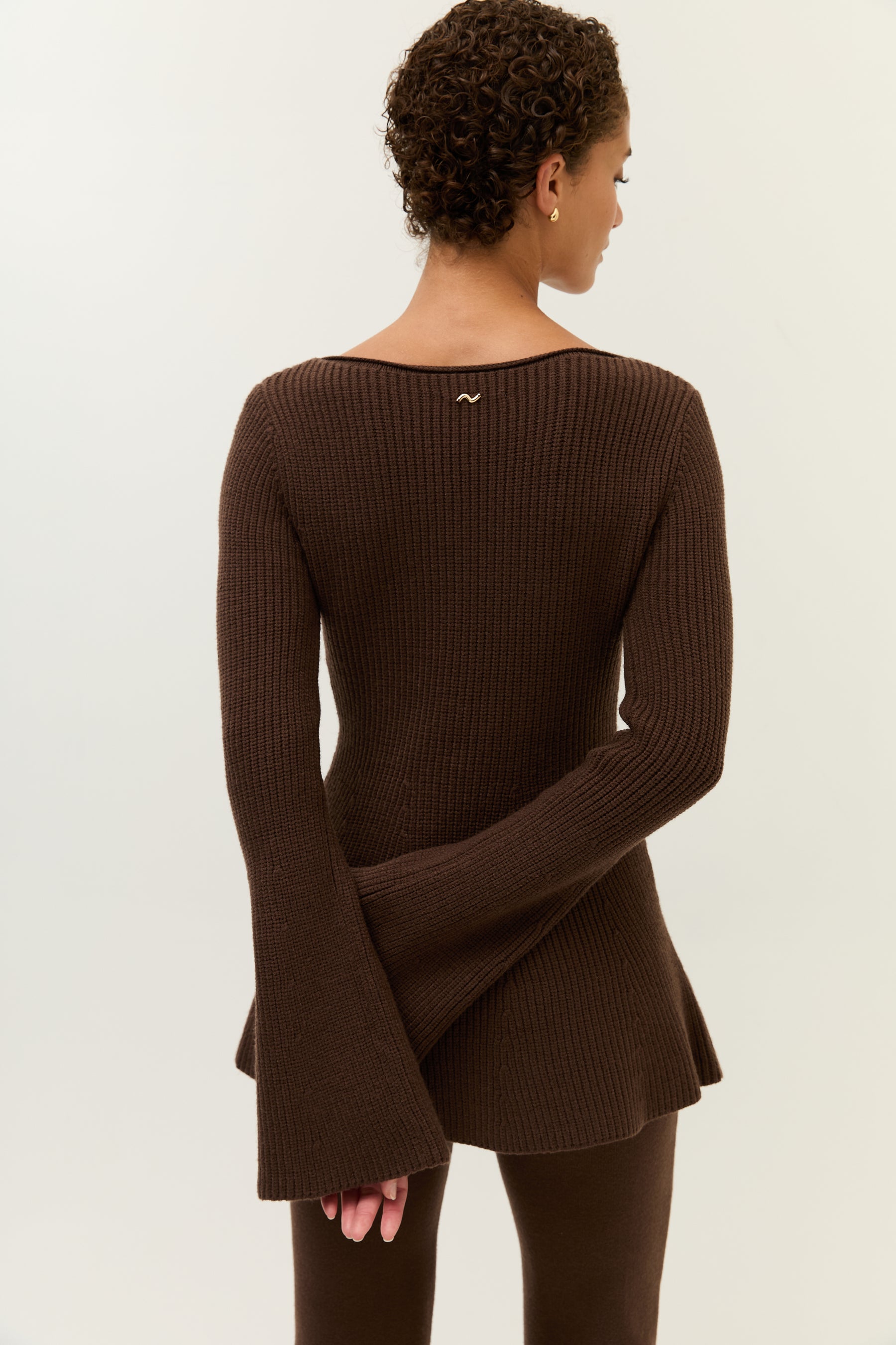 Frau trägt ein lockeres, zweiteiliges Set aus strukturiertem Bouclé-Stoff in dunklem Schokobraun. Das Set besteht aus einem langärmeligen, weiten Pullover mit Rundhalsausschnitt und einer passenden, weiten Hose mit geradem Bein. Sie kombiniert das Outfit mit goldenen Creolen und schwarzen Loafern. Der Look wirkt gemütlich und entspannt.