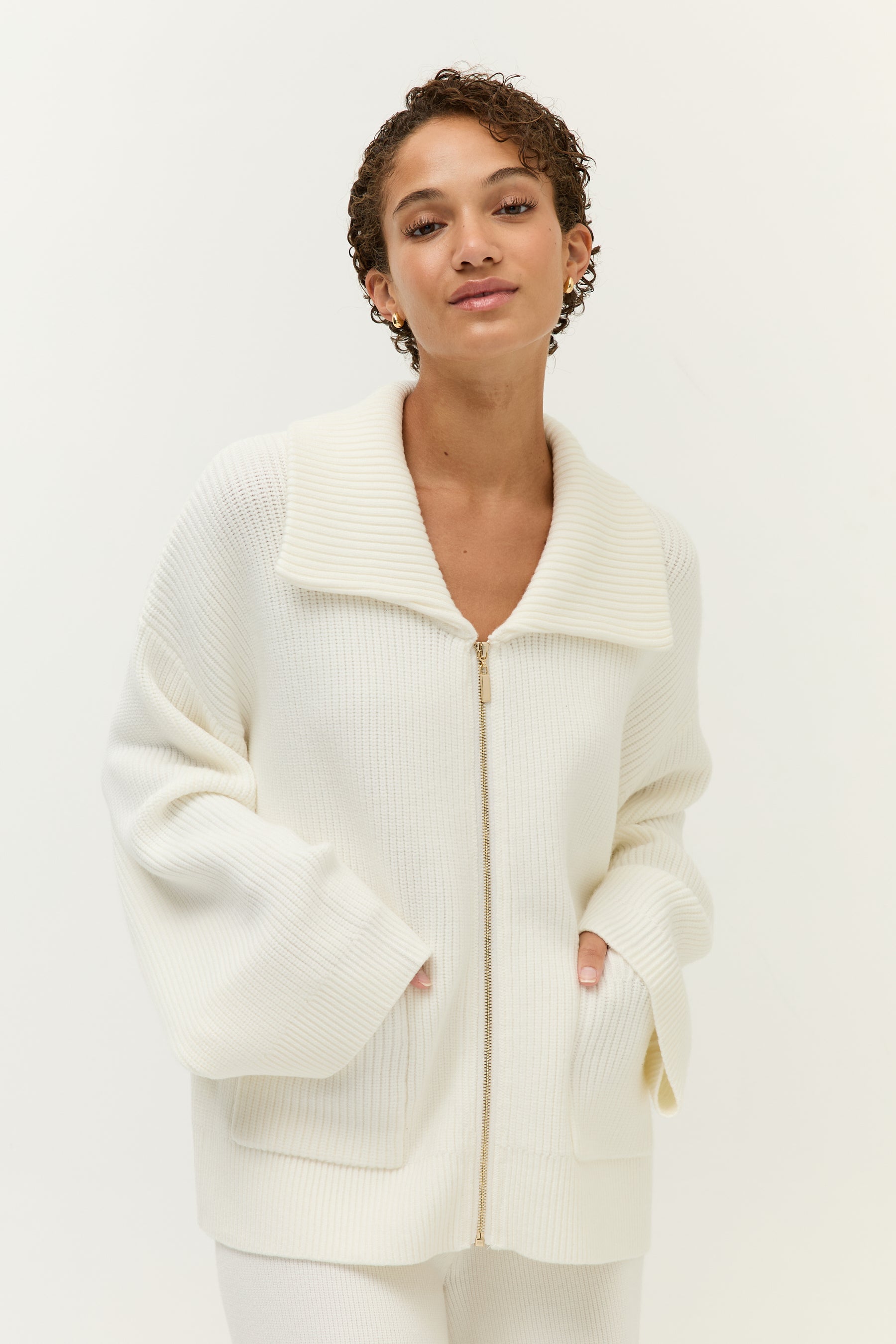 Look 2 – Strickset in Off-White (Bilder 7–12):
Modell trägt ein zweiteiliges Set aus weichem, geripptem Strick in Off-White, bestehend aus einem langärmeligen Zip-Cardigan mit breitem Kragen, goldfarbenem Reißverschluss und zwei großen aufgesetzten Taschen sowie einer weit geschnittenen Hose mit fließendem Fall. Auf der Rückseite der Jacke ist ein kleines, goldfarbenes Lezu-Logo unter dem Kragen platziert. Styling mit beigen Slippern.