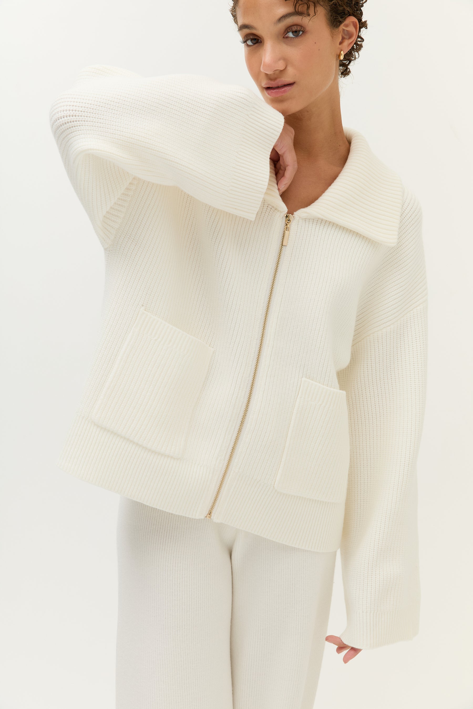 Look 2 – Strickset in Off-White (Bilder 7–12):
Modell trägt ein zweiteiliges Set aus weichem, geripptem Strick in Off-White, bestehend aus einem langärmeligen Zip-Cardigan mit breitem Kragen, goldfarbenem Reißverschluss und zwei großen aufgesetzten Taschen sowie einer weit geschnittenen Hose mit fließendem Fall. Auf der Rückseite der Jacke ist ein kleines, goldfarbenes Lezu-Logo unter dem Kragen platziert. Styling mit beigen Slippern.