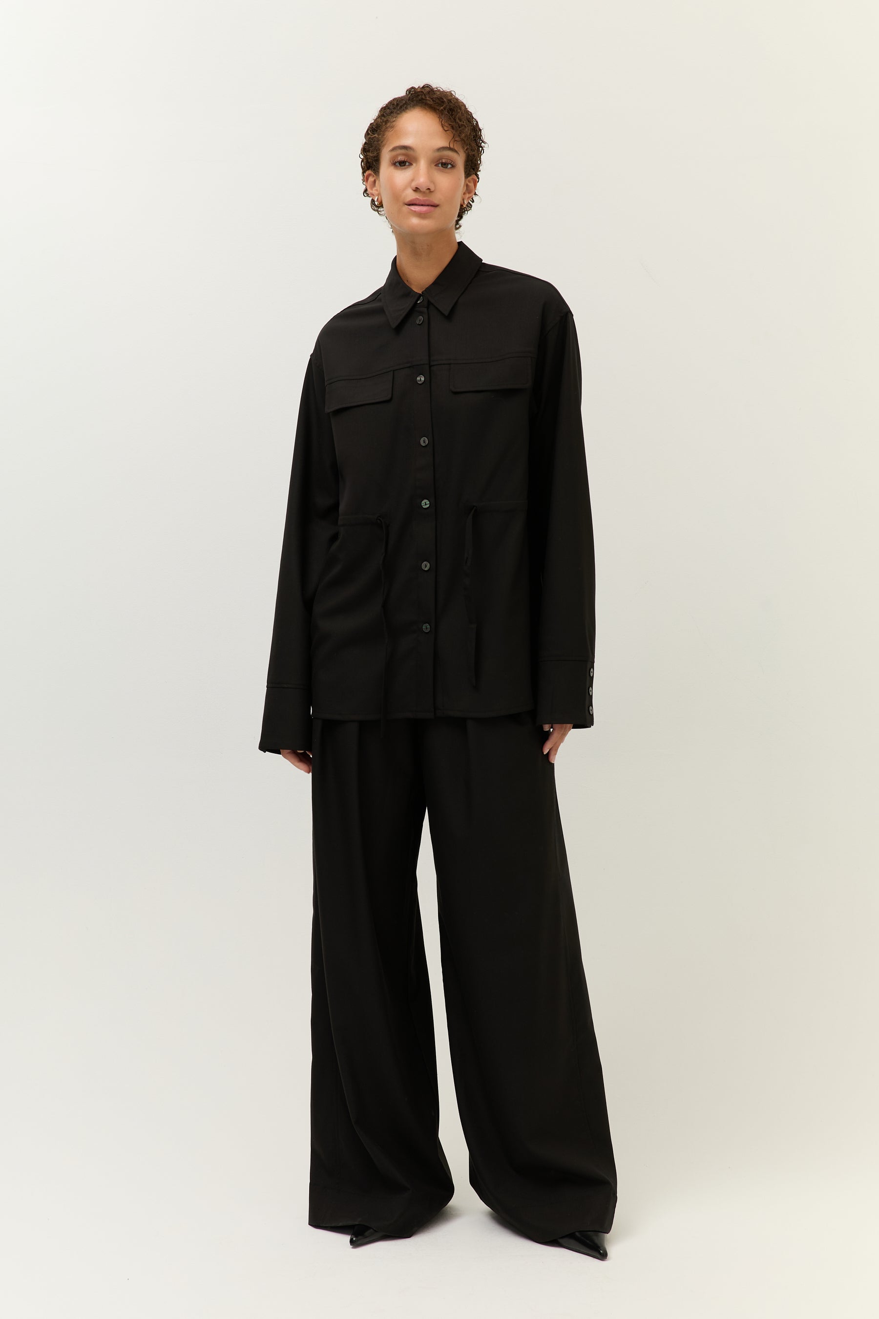Frontalansicht, offen getragen
Model trägt schwarzen Zweiteiler mit gerader Hemdjacke, Brusttaschen und weiter Hose.