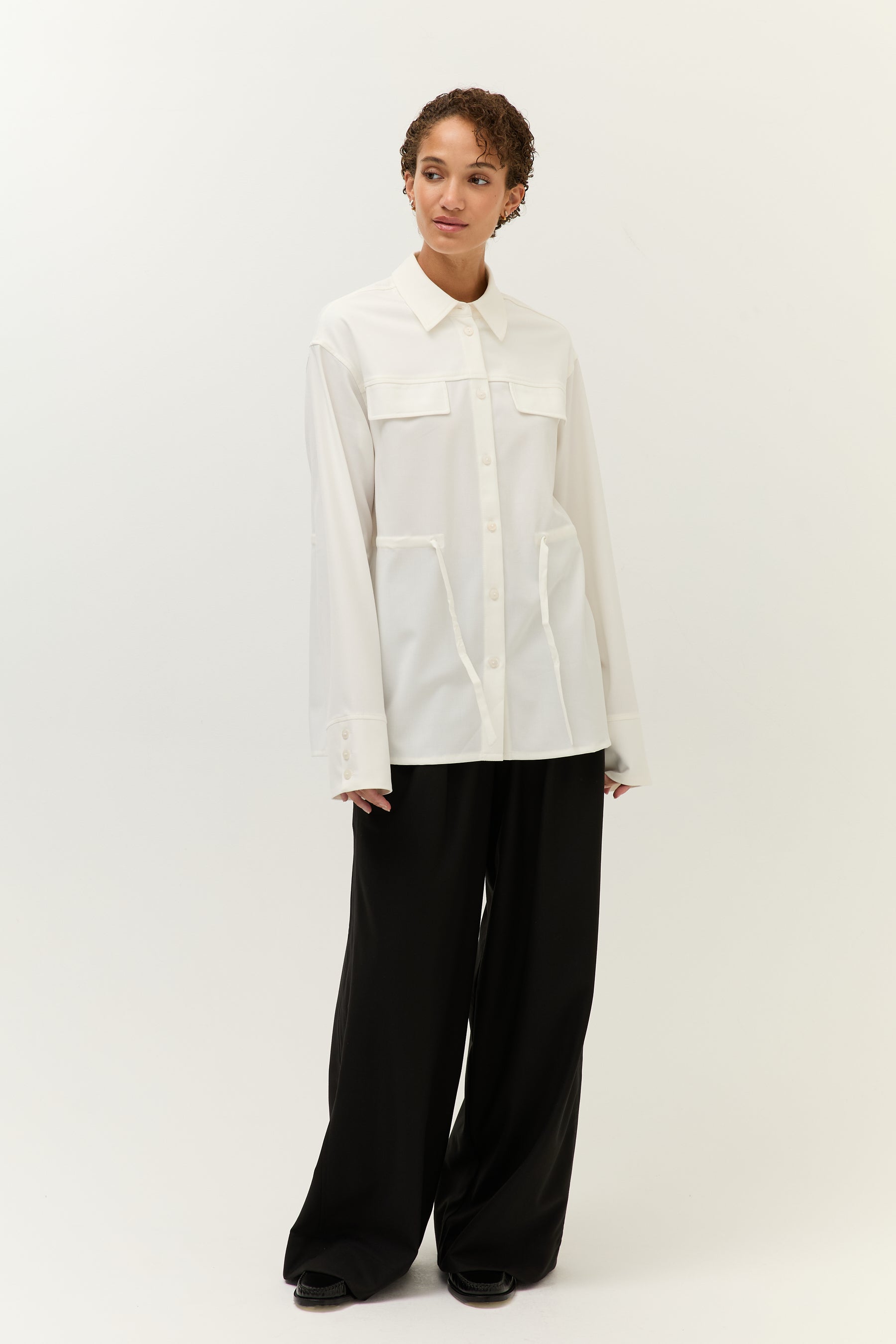 Frau steht leicht seitlich gedreht vor hellem Hintergrund und trägt eine weiße, locker geschnittene Bluse mit aufgesetzten Brusttaschen, durchgehender Knopfleiste und Bindegürtel auf Hüfthöhe. Dazu kombiniert sie eine weite schwarze Hose.