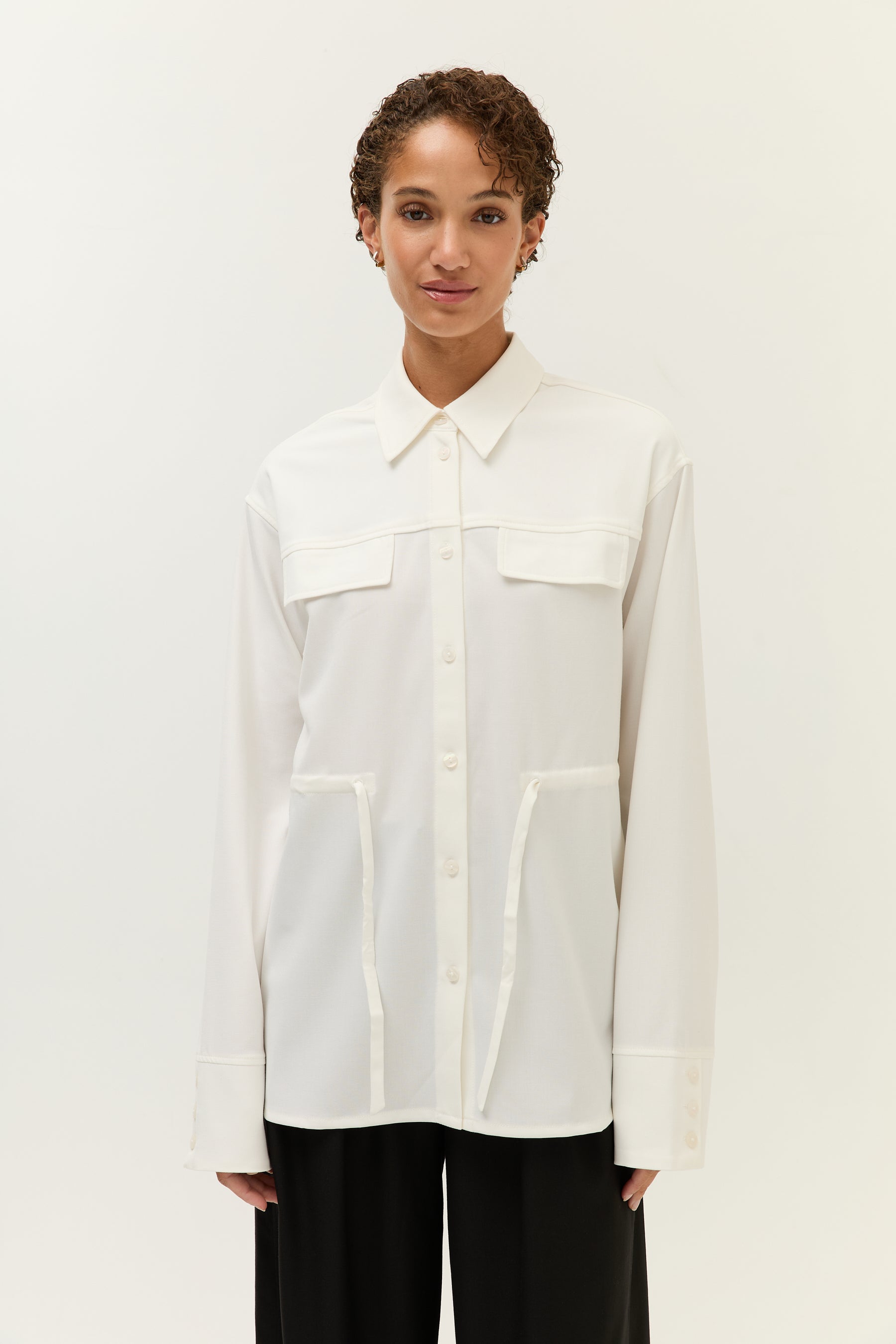 Frontalaufnahme der Frau mit derselben weißen Bluse. Gut sichtbar sind der klassische Kragen, die Knopfleiste und die Bindebänder, die locker aus den seitlichen Taschen hängen.