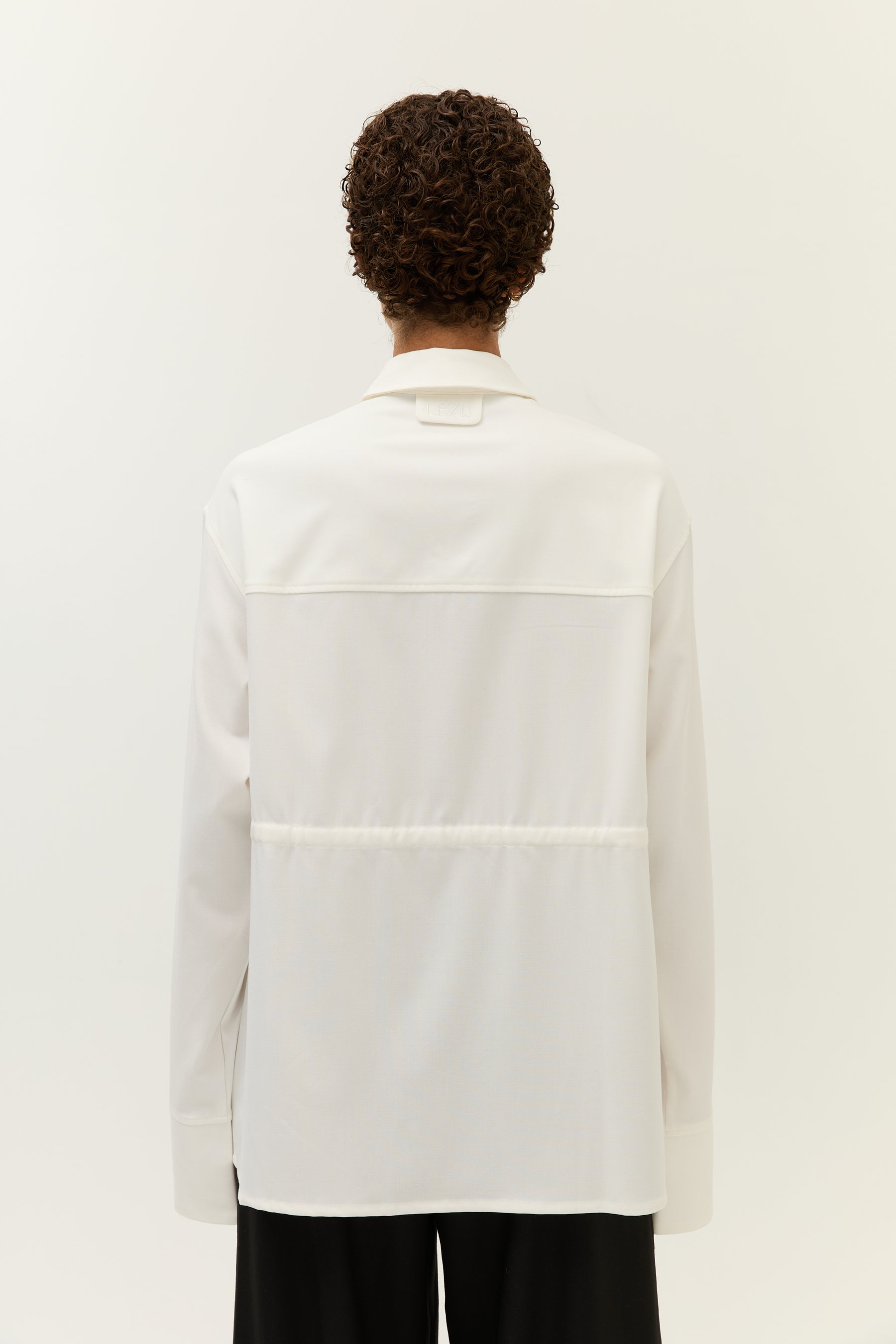 Rückansicht der Frau mit geschlossener weißer Bluse. Im Nackenbereich ist ein kleines, aufgenähtes Label mit „LEZU“-Prägung zu erkennen.