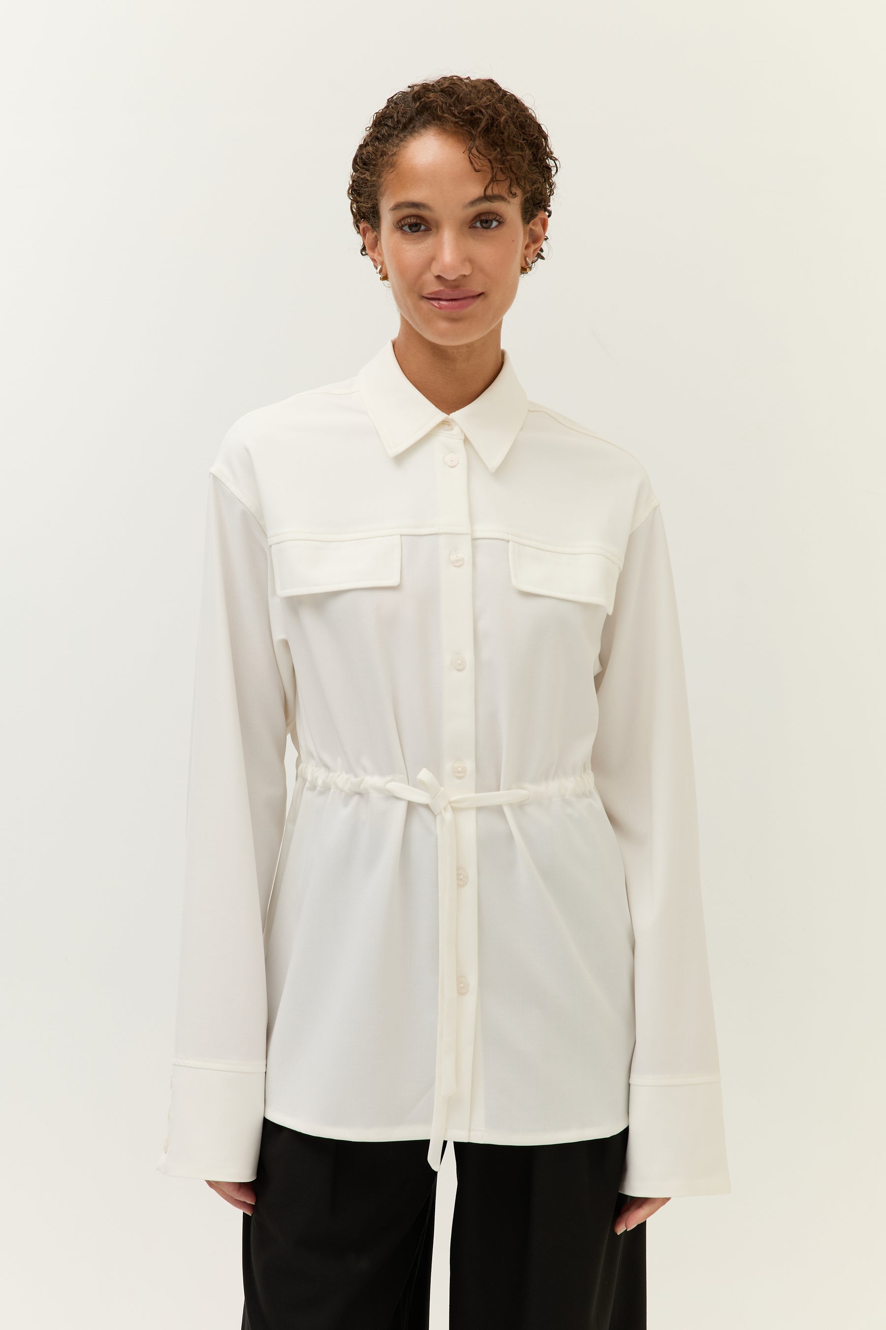 Frau steht frontal vor hellem Hintergrund und trägt ein weißes Hemdblusenshirt mit aufgesetzten Brusttaschen, durchgehender Knopfleiste und Tunnelzug in der Taille, kombiniert mit schwarzer Hose.