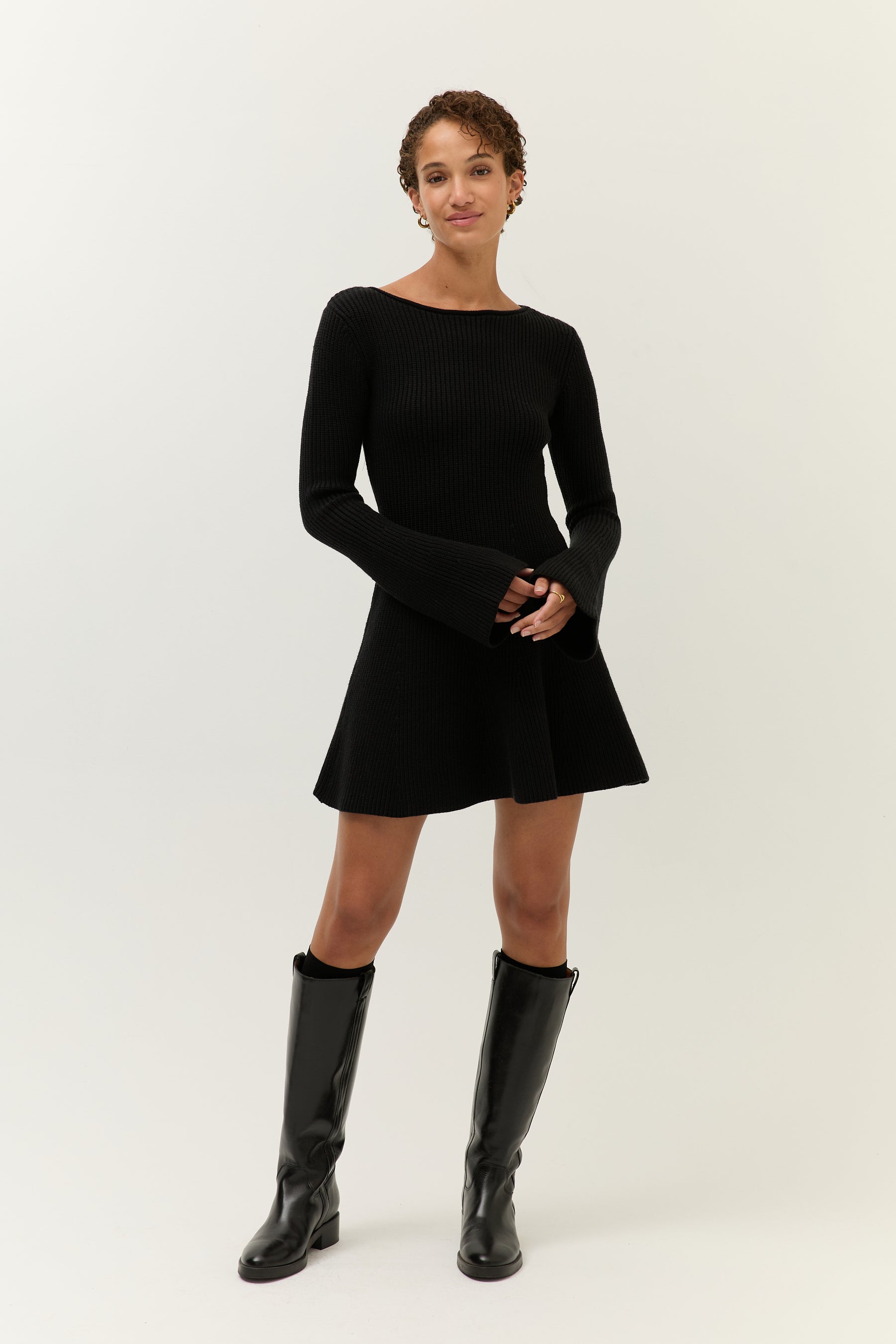 Model in einem schwarzen, gerippten Strickkleid mit ausgestelltem Rock und langen, leicht ausgestellten Ärmeln. Sie trägt kniehohe schwarze Lederstiefel und goldene Ohrringe. Blick frontal zur Kamera mit leicht geneigtem Kopf.