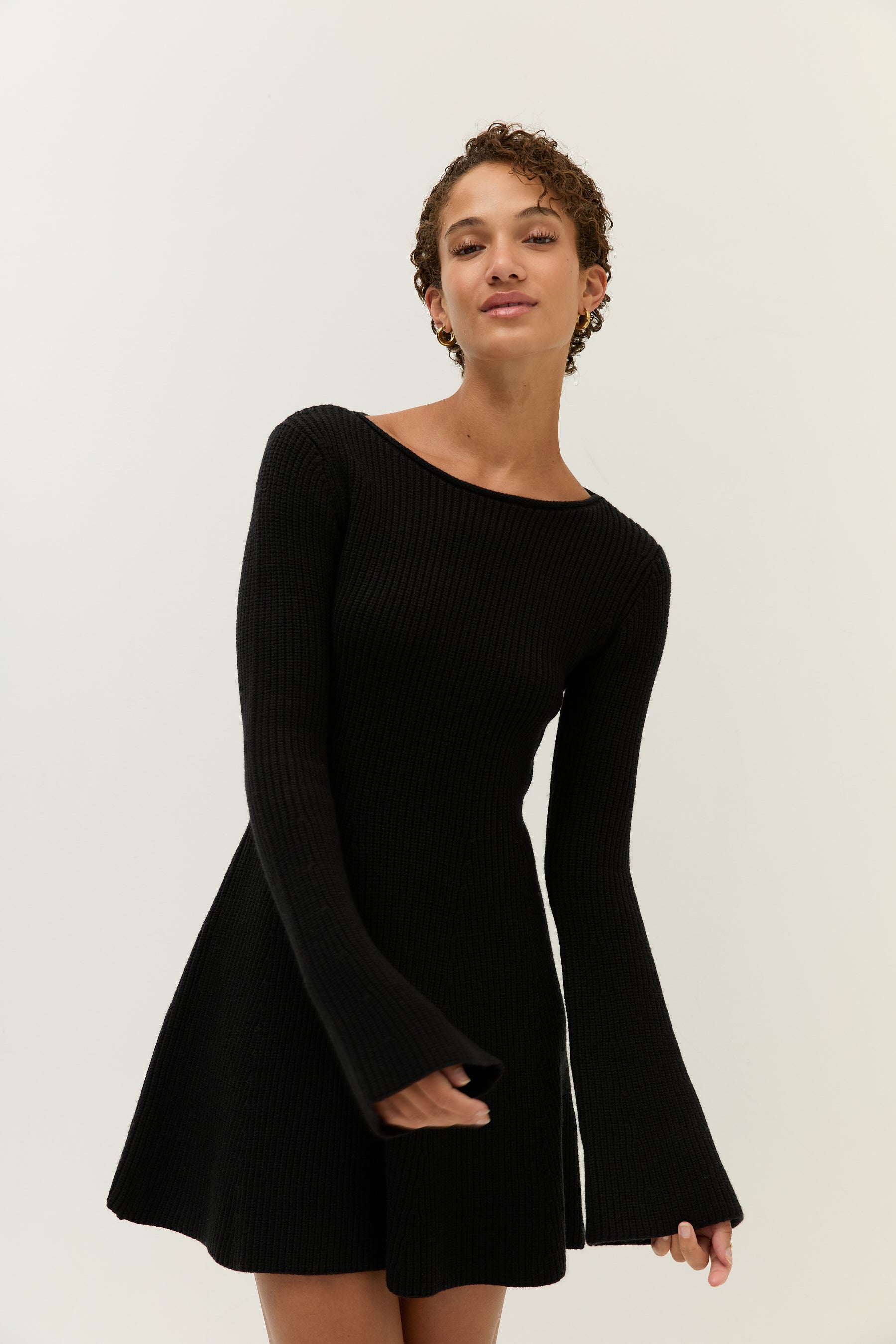 Nahaufnahme des Oberkörpers im schwarzen Strickkleid mit weitem Rundhalsausschnitt. Das Model blickt selbstbewusst in die Kamera und zeigt die langen, ausgestellten Ärmel.