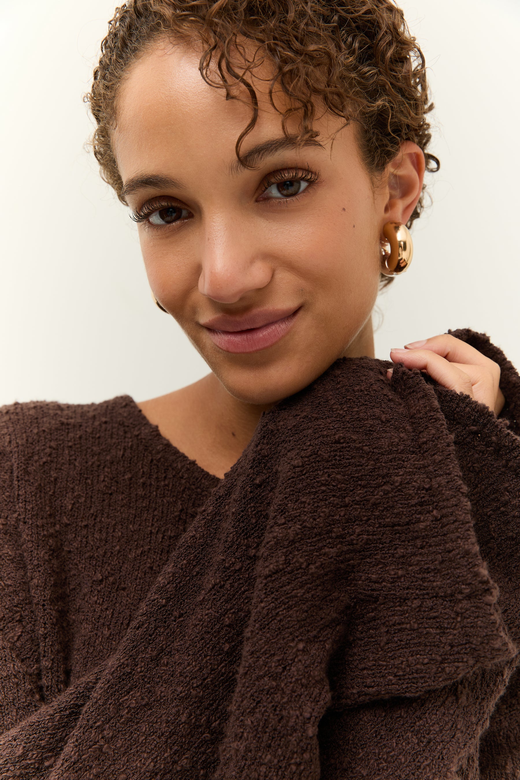 Nahaufnahme des Gesichts mit Fokus auf die großen, goldenen Ohrringe und die Bouclé-Struktur am Kragen.