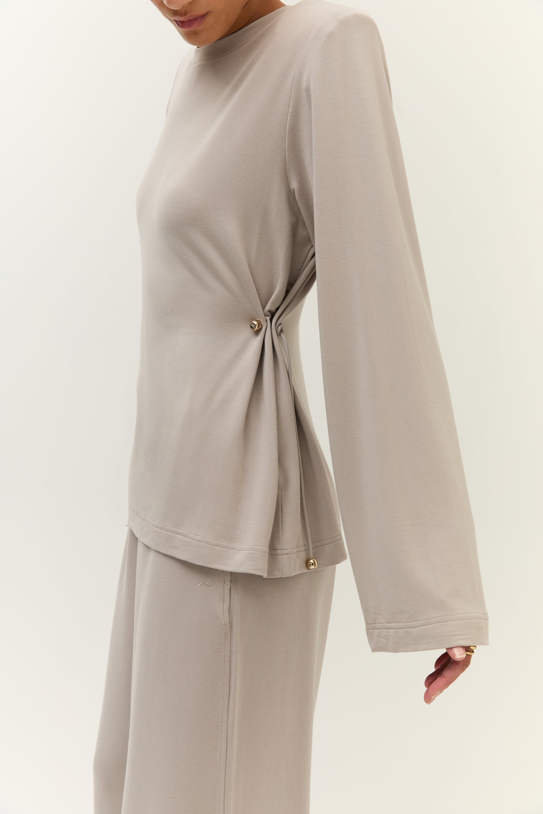 Seitliche Ansicht des Oberteils mit Raffungsdetail
Seitliche Ansicht eines taupefarbenen Oberteils mit lockerem Schnitt, weitem Rundhalsausschnitt und Tunnelzug mit goldenen Endkappen an der Taille.