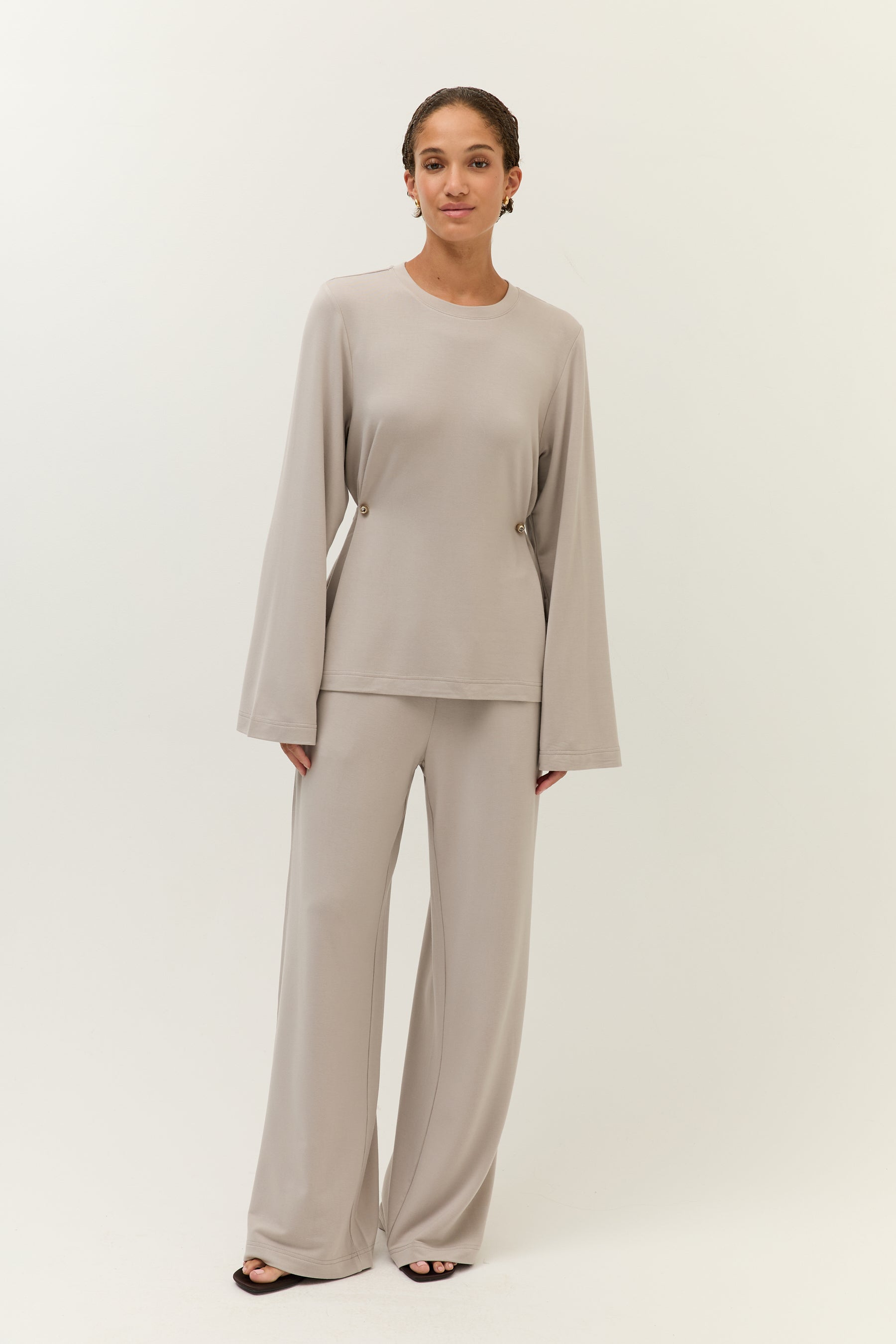 Das Model trägt ein monochromes Set in Taupe mit lockerem, gerafftem Oberteil und weit geschnittener Hose, kombiniert mit schwarzen offenen Schuhen.