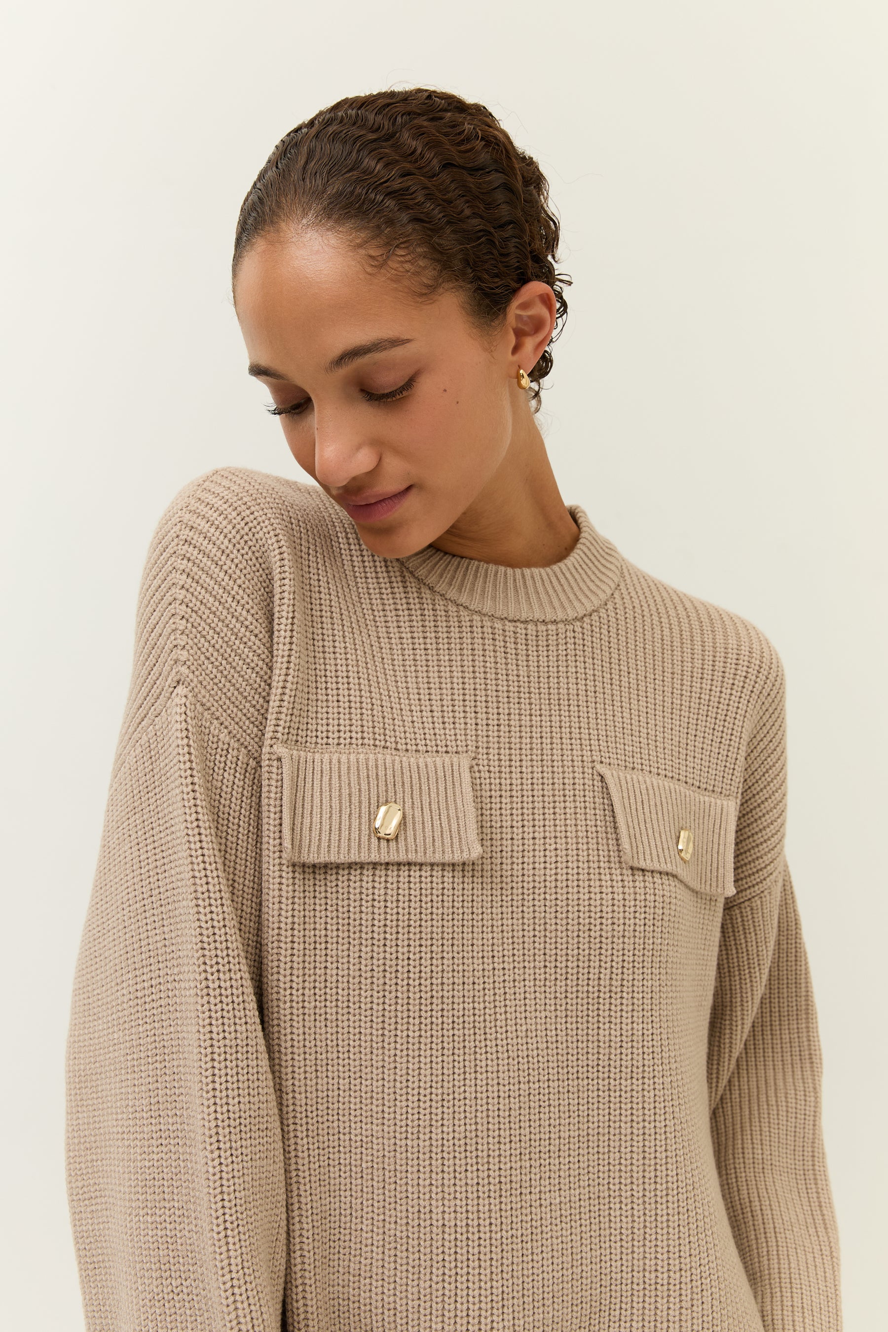 Portrait mit Blick nach unten
Model im sandfarbenen Strickkleid mit zwei Taschen vorne, die mit goldenen Zierknöpfen versehen sind.