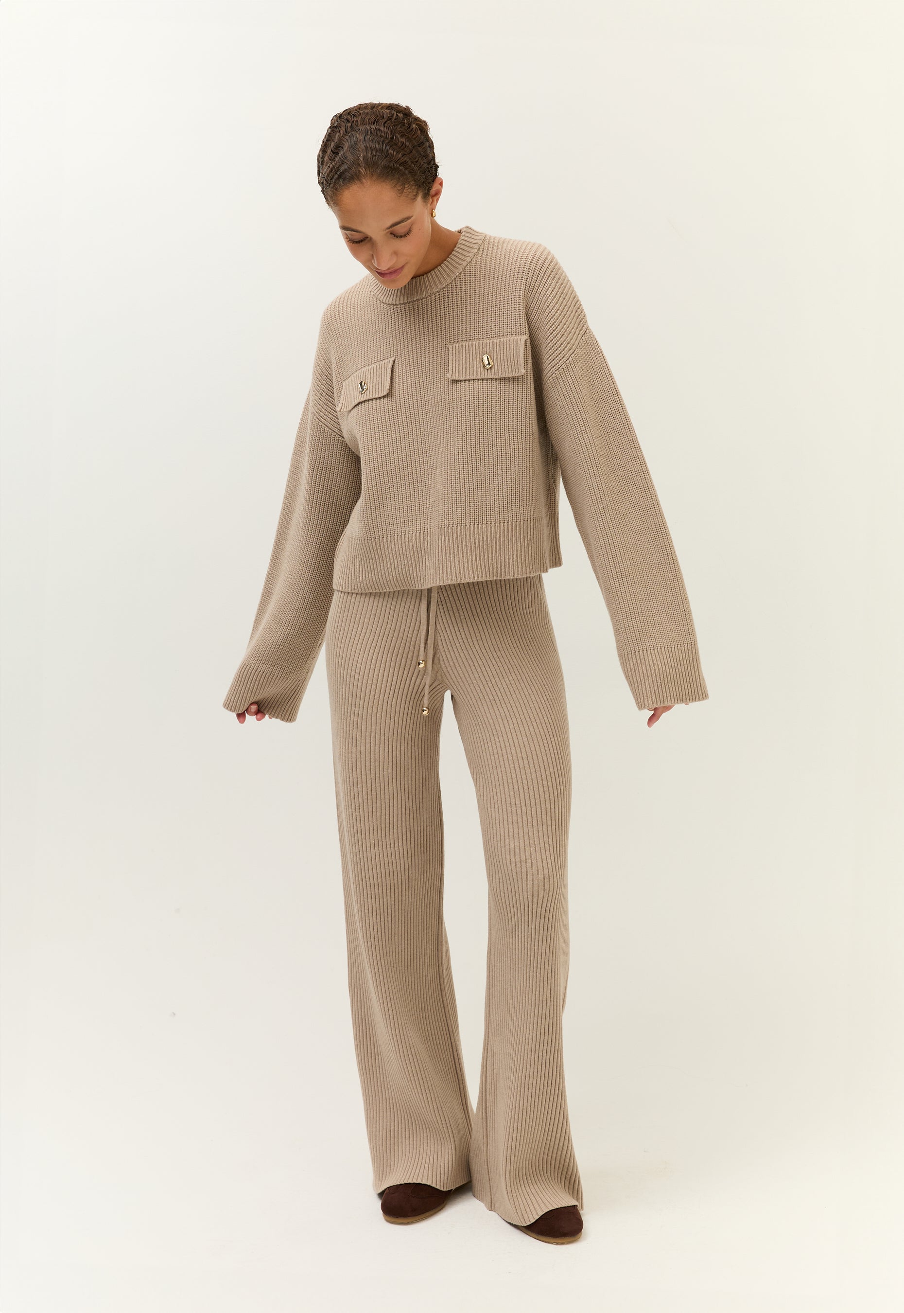 Frau in einem beigen, grob gestrickten Zweiteiler mit weitem Pulli und Hose, steht mit gesenktem Blick und gespreizten Armen vor hellem Hintergrund.