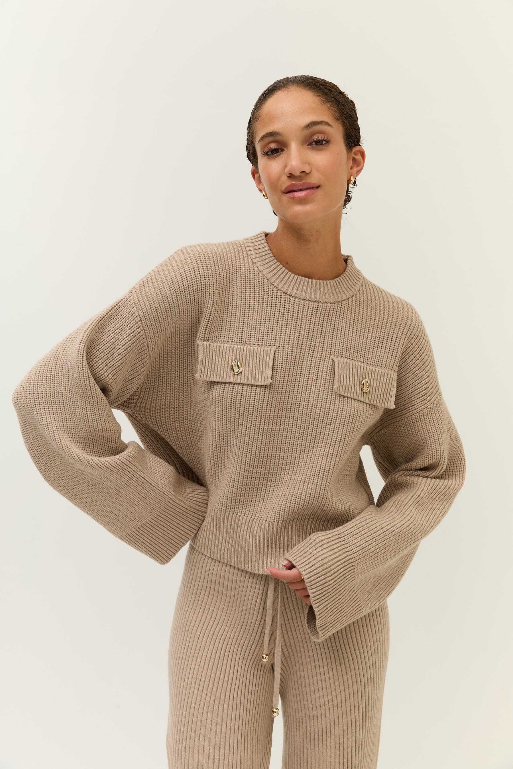 Porträt einer Frau in beige, die Hände in die Taille gestützt, der Strickpullover mit zwei Brusttaschen samt goldenen Knöpfen.