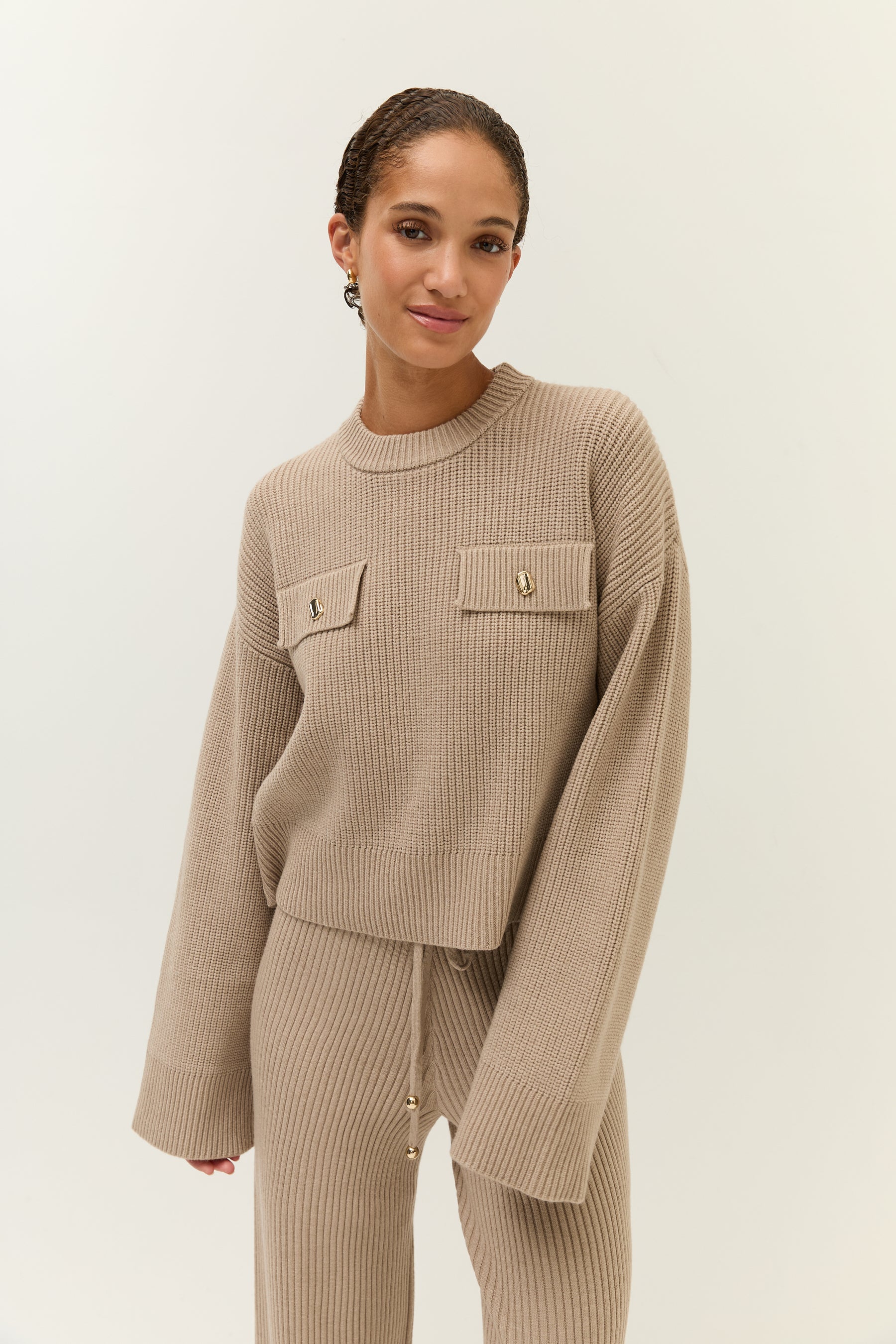 Frau im beigefarbenen Strick-Zweiteiler, frontal zur Kamera, mit lockerem Gesichtsausdruck und locker fallenden Ärmeln.