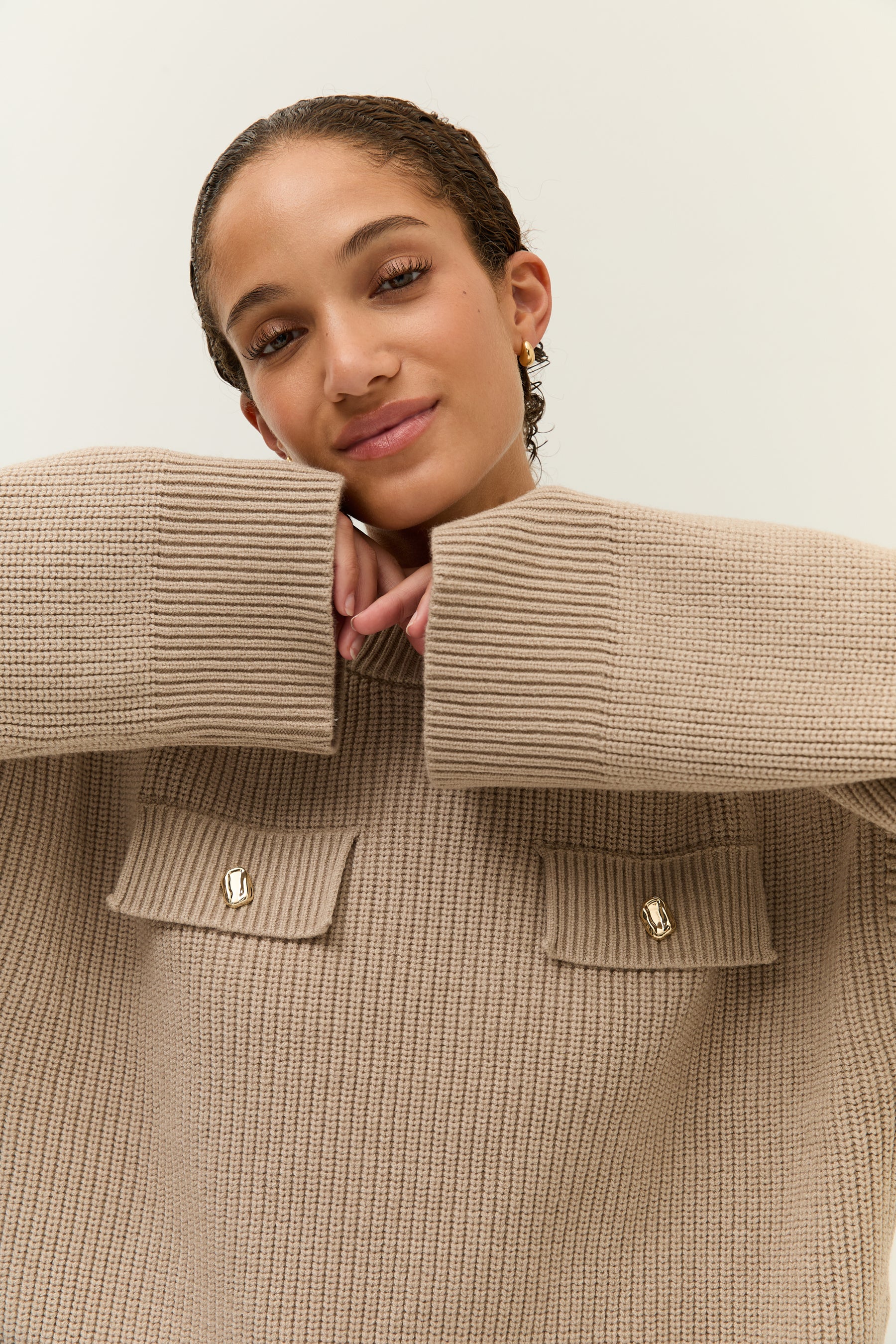 Nahaufnahme des Pullovers mit Fokus auf die Brusttaschen mit goldenen Knöpfen; die Frau stützt ihr Kinn auf die hochgezogenen Ärmel.
