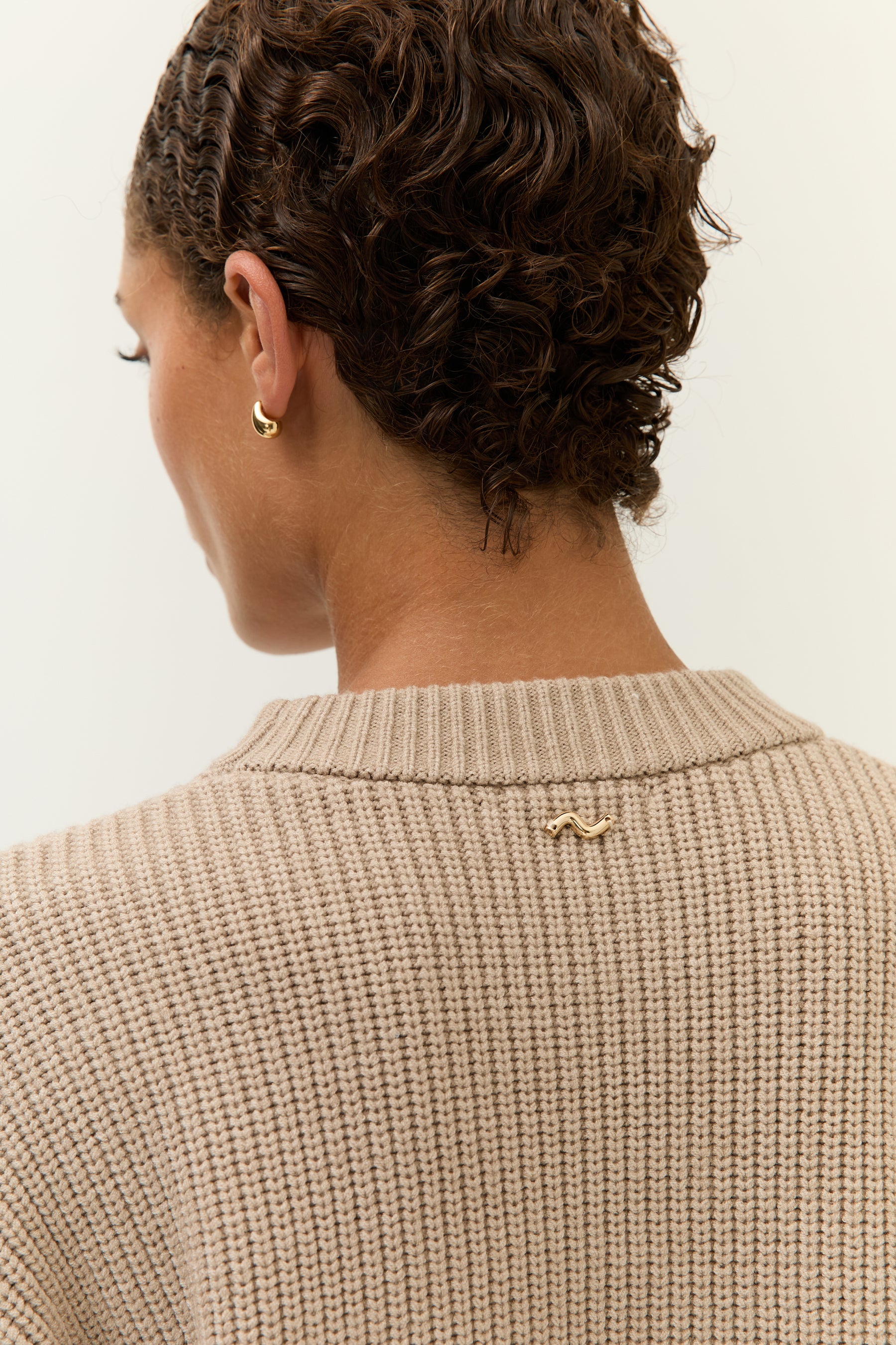 Detailaufnahme des Nackens der Frau; zu sehen ist das goldene Wellen-Logo auf dem Rücken des beigefarbenen Strickpullovers.